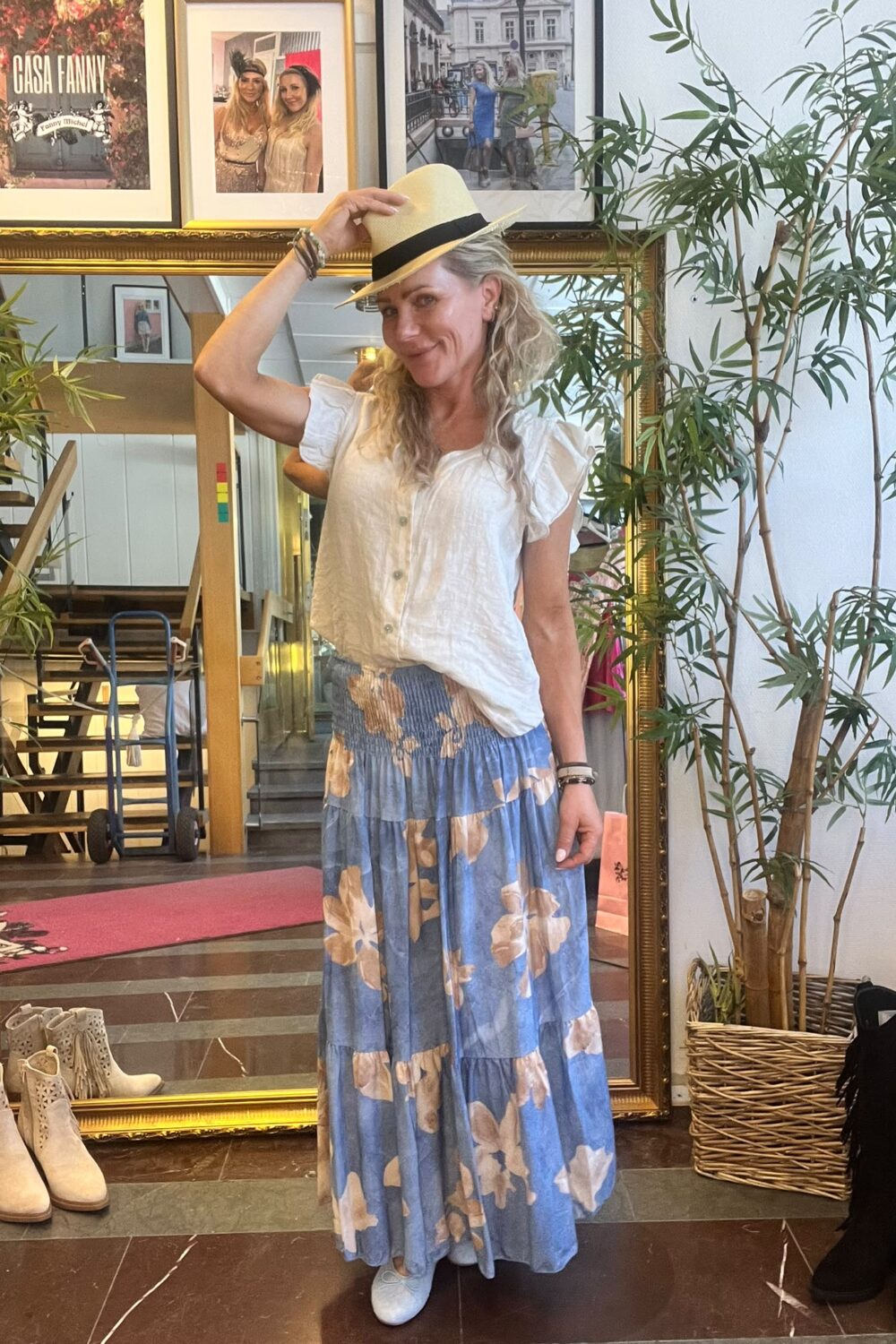 Kvinna står och lutar sig lätt fram, håller i brättet på en ljus hatt och ler. Hon bär vit blus och blå långkjol med blommor, framför en stor spegel och växter; väggkonst med texten ”CASA FANNY”.