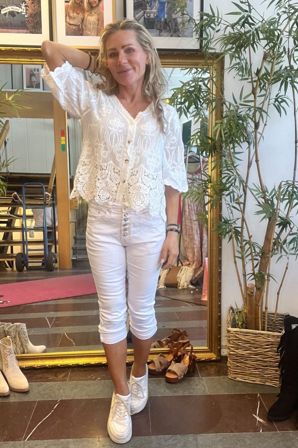Leende kvinna i vit, broderad blus och vita capribyxor poserar med ena armen bakom huvudet. Hon står framför en stor guldramsspegel i en butiksmiljö med skor på golvet, växt i korg och inramade foton.
