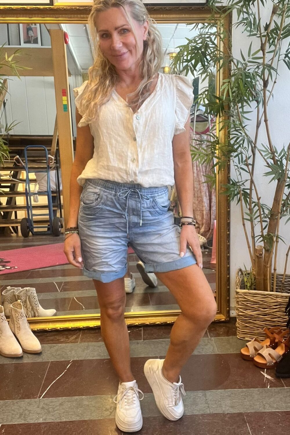 Kvinna står och ler mot kameran med ena knät lätt böjt, klädd i vit blus, blå shorts och vita sneakers. Hon poserar framför en stor guldramsspegel i en butik med skor och en krukväxt.