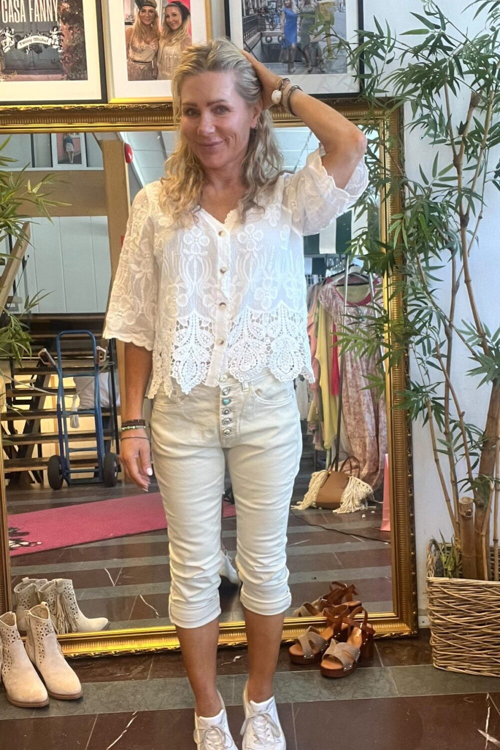 Kvinna i vit spetsblus och ljusa capribyxor poserar framför en stor guldramsspegel, med ena handen i håret och ett leende. I bakgrunden syns växter, skor på golvet och inramade foton; text: “CASA FANNY”.