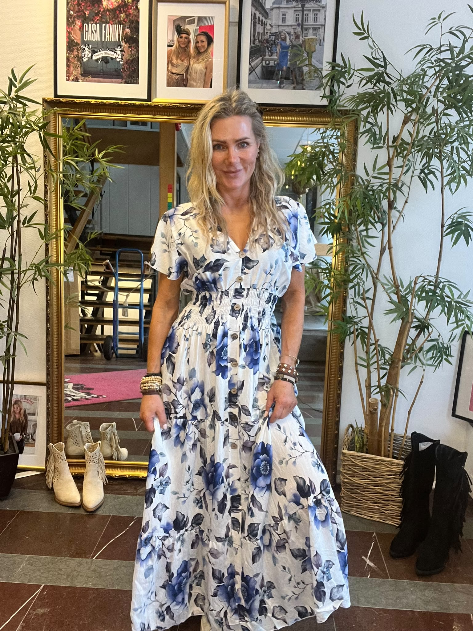 Blond kvinna står och ler framför en stor guldramad spegel och håller ut sin vitblå blommiga klänning. Runt henne syns gröna krukväxter, skor på golvet och tre inramade foton ovanför, varav ett med texten “CASA FANNY”.