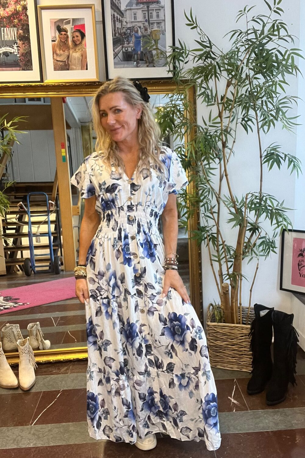 Blond kvinna i vit maxiklänning med blå blomster står och ler framför en stor guldramsspegel i en butiksmiljö. Runt henne syns gröna krukväxter, boots på golvet och inramade foton; en skylt säger “CASA FANNY”.