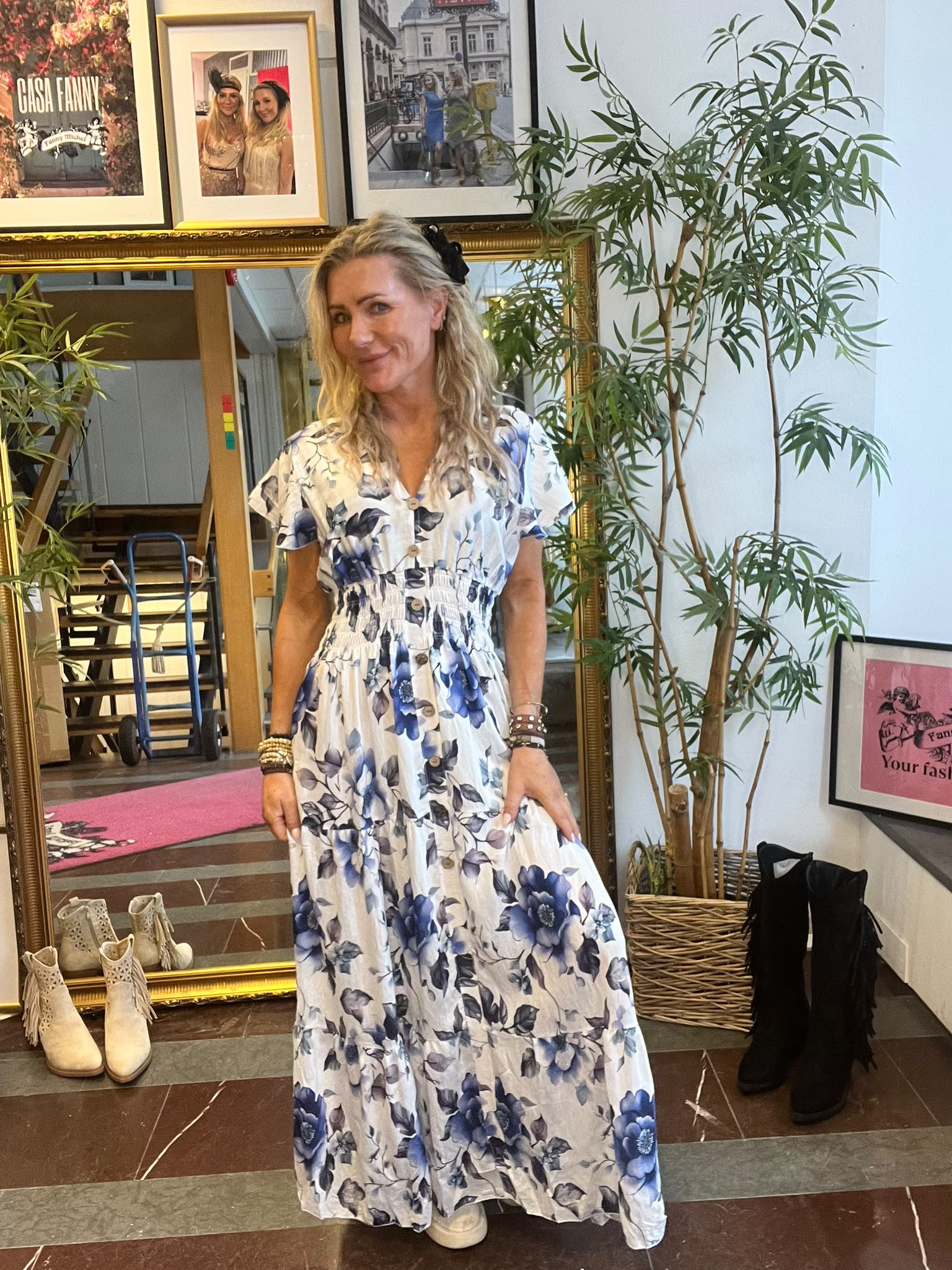 Blond kvinna i vit maxiklänning med blå blomster står och ler framför en stor guldramsspegel i en butiksmiljö. Runt henne syns gröna krukväxter, boots på golvet och inramade foton; en skylt säger “CASA FANNY”.