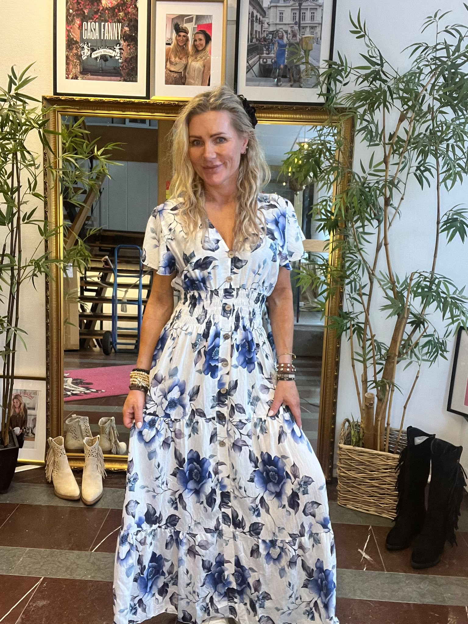 Blond kvinna står och ler i en butiksmiljö, klädd i vit lång klänning med blå blommor och knappar. Hon håller lätt i kjolen. Bakom syns guldramsspegel, gröna växter, skor och inramade foton; text: “CASA FANNY”.
