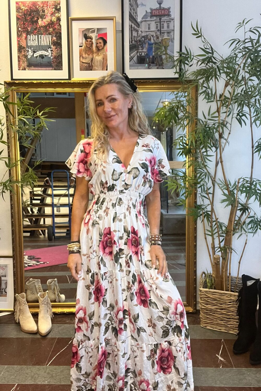 Leende kvinna poserar i en vit klänning med stora rosa blommor framför en guldramad spegel i en butik. Runt henne syns krukväxter, boots på golvet och inramade foton; en tavla har texten “CASA FANNY”.