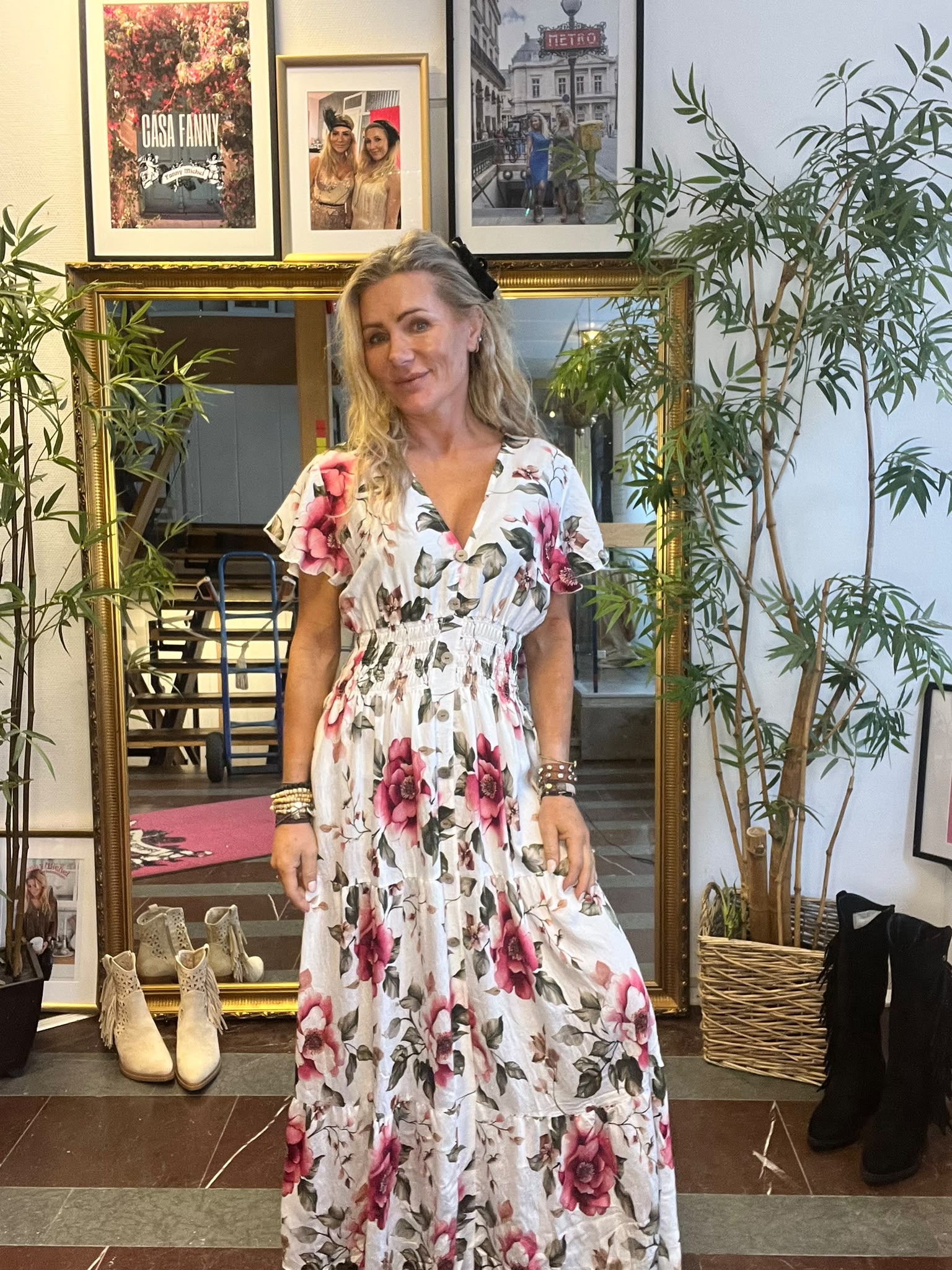 Leende kvinna poserar i en vit klänning med stora rosa blommor framför en guldramad spegel i en butik. Runt henne syns krukväxter, boots på golvet och inramade foton; en tavla har texten “CASA FANNY”.