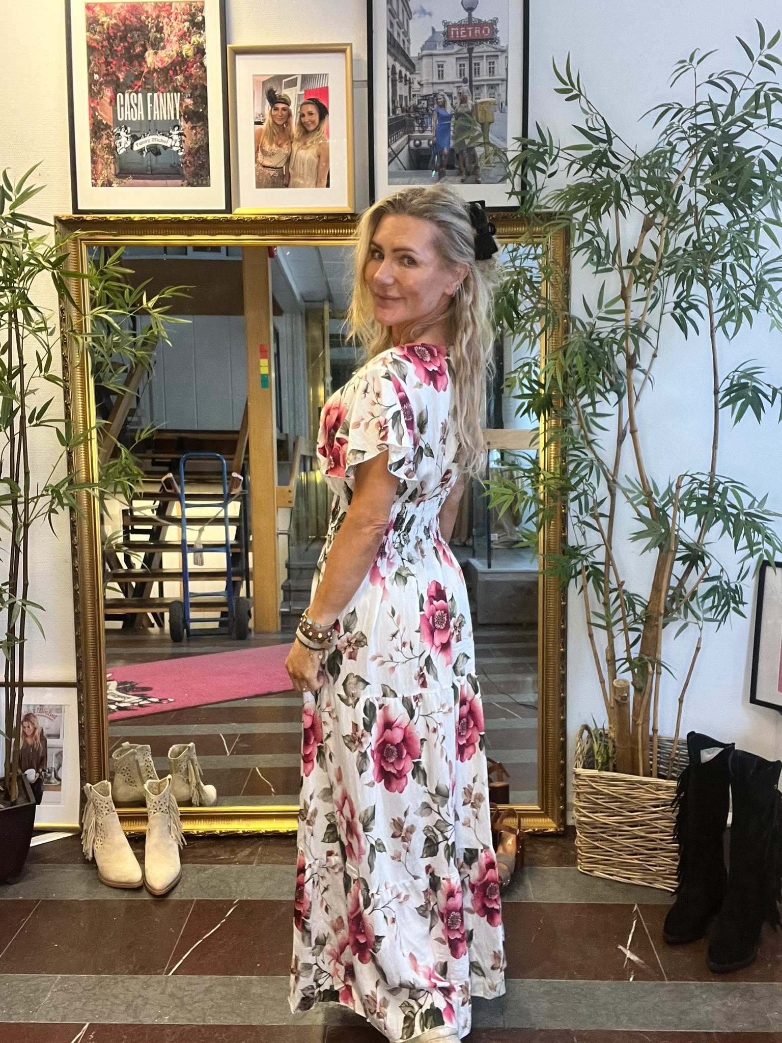 Kvinna i vit, blommig långklänning står och ler över axeln i en butik, framför en stor guldramsspegel. Runt henne finns växter, skor på golvet och väggfoton med texten ”CASA FANNY” och ”METRO”.