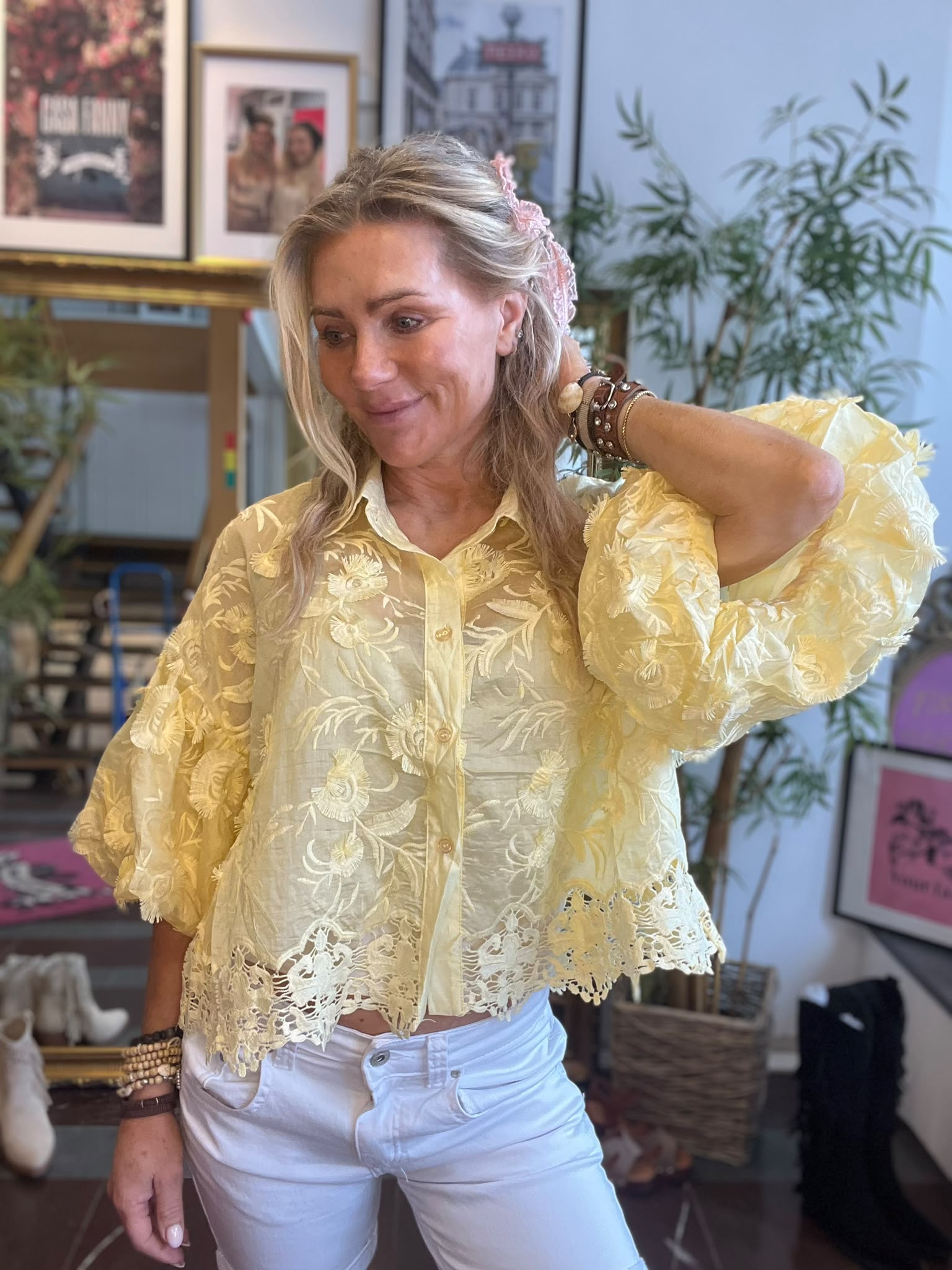 Blond kvinna i ljusgul, broderad blus och vita byxor står inomhus och lyfter handen till håret med ett svagt leende. I bakgrunden syns inramade affischer och en grön växt.