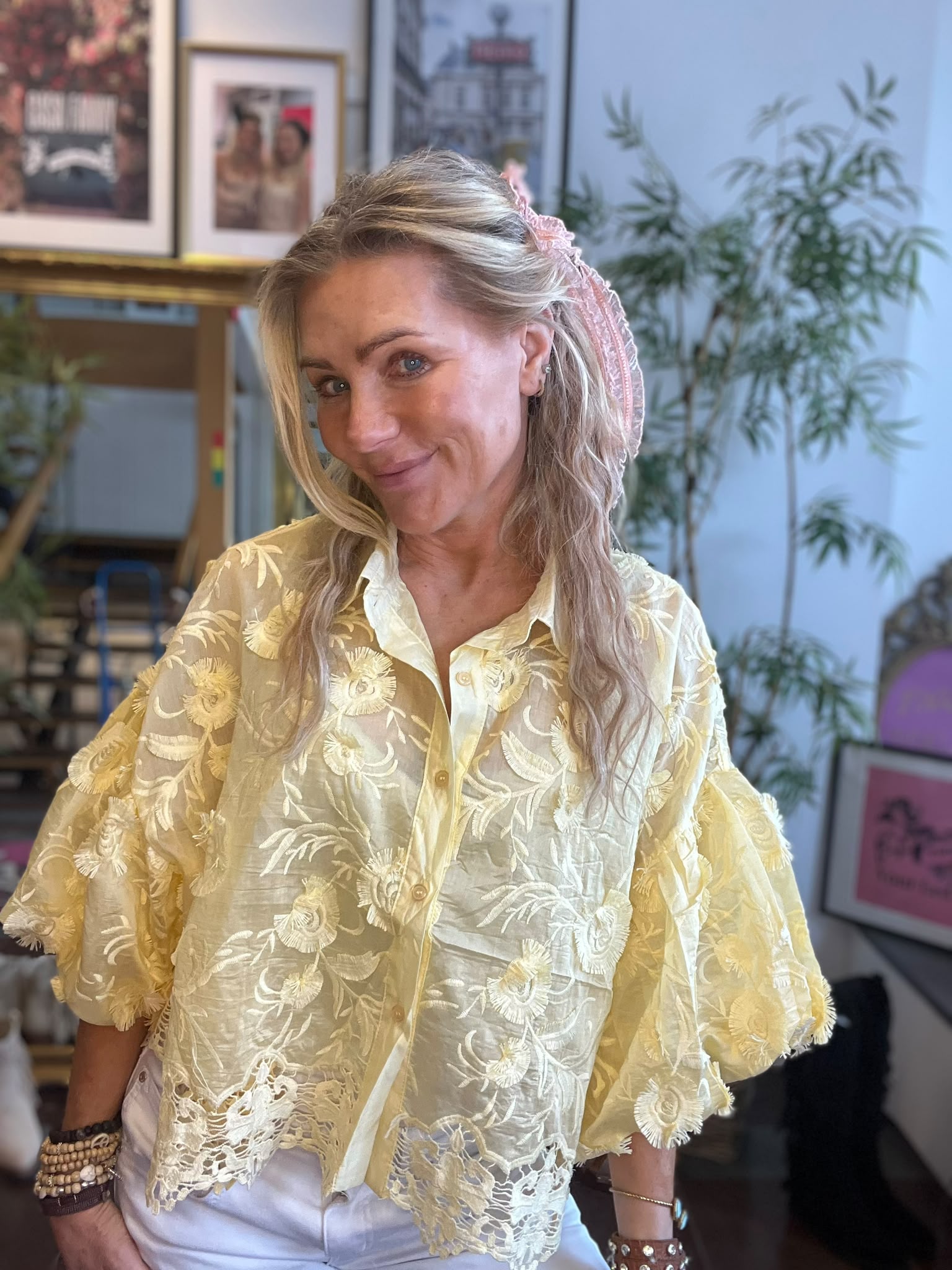 Leende kvinna poserar inomhus med huvudet lätt på sned och handen på höften. Hon har långt blont hår med rosa hårband och bär en gul broderad blus. I bakgrunden syns växt och inramade bilder.