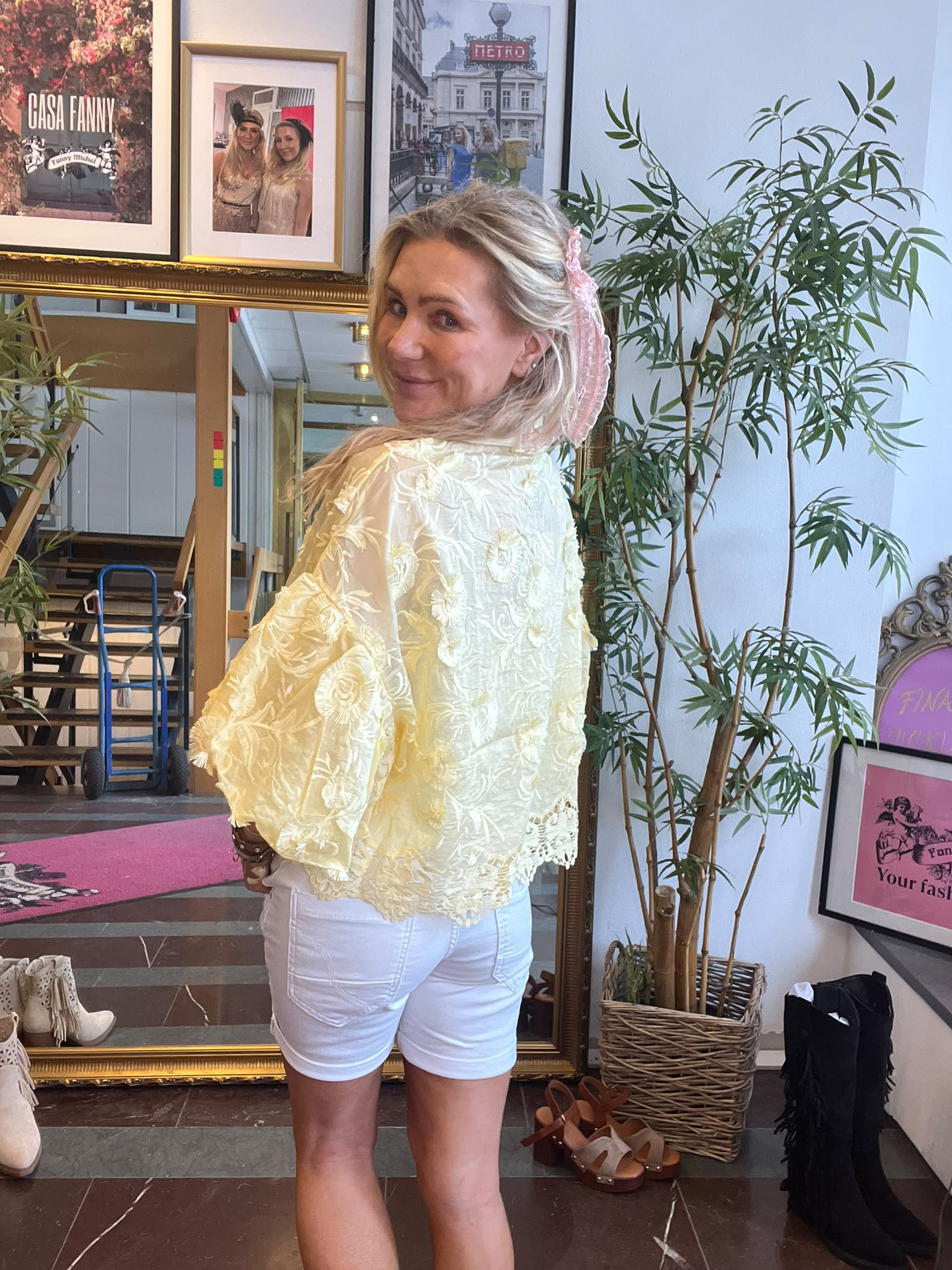 Leende kvinna vänder sig mot kameran med en rosa håraccessoar, iklädd ljusgul spetsblus och vita shorts. Hon står framför en stor guldramsspegel i en butik med växter och inramade affischer, bland annat “CASA FANNY” och “METRO”.