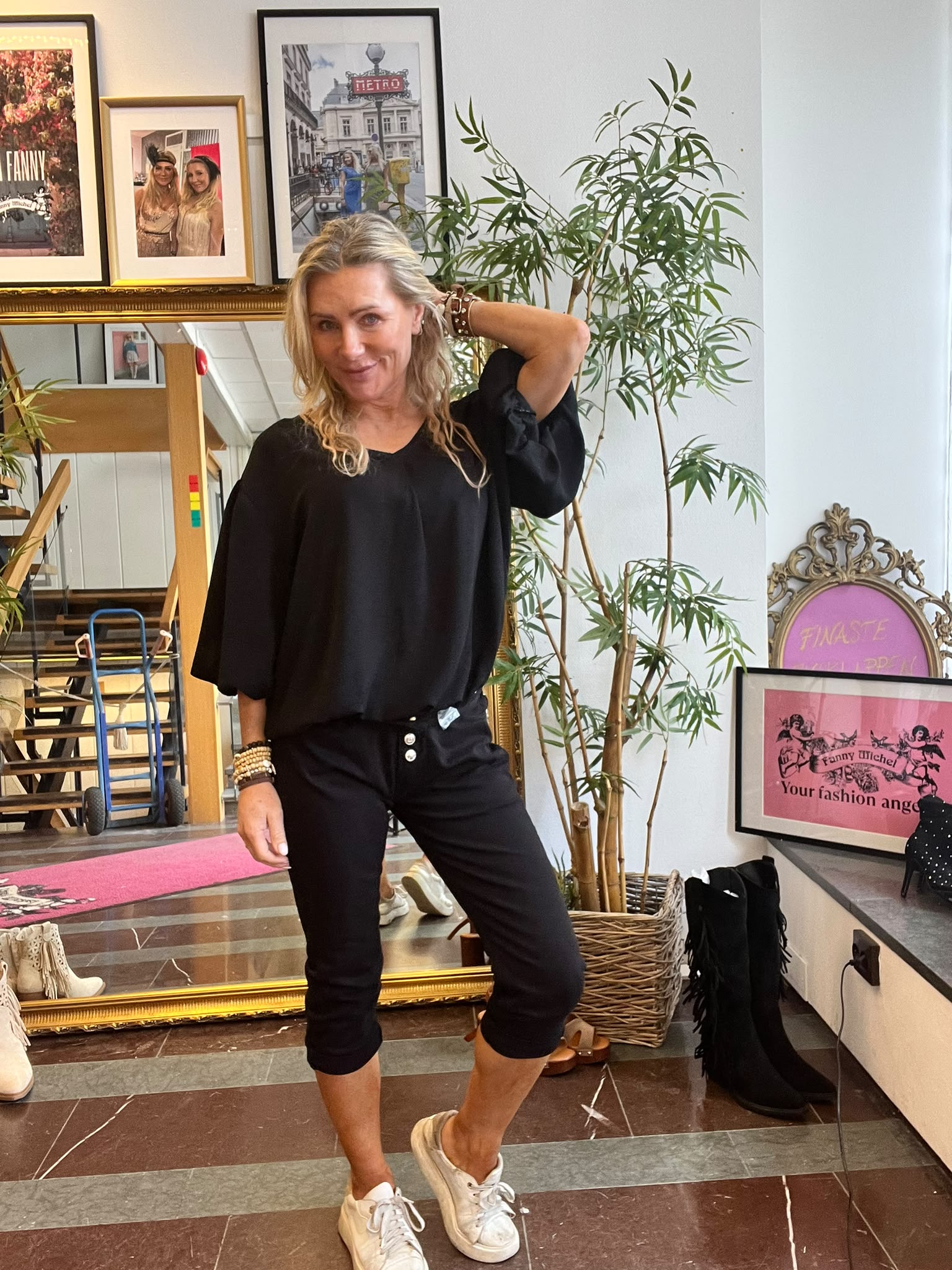 Kvinna i svart outfit poserar framför en stor guldspegel med handen bakom huvudet och ena benet fram. Hon står i en butikslik miljö med växt, skor och inramade posters, bland annat texten “Fanny” och “Your fashion ange”.