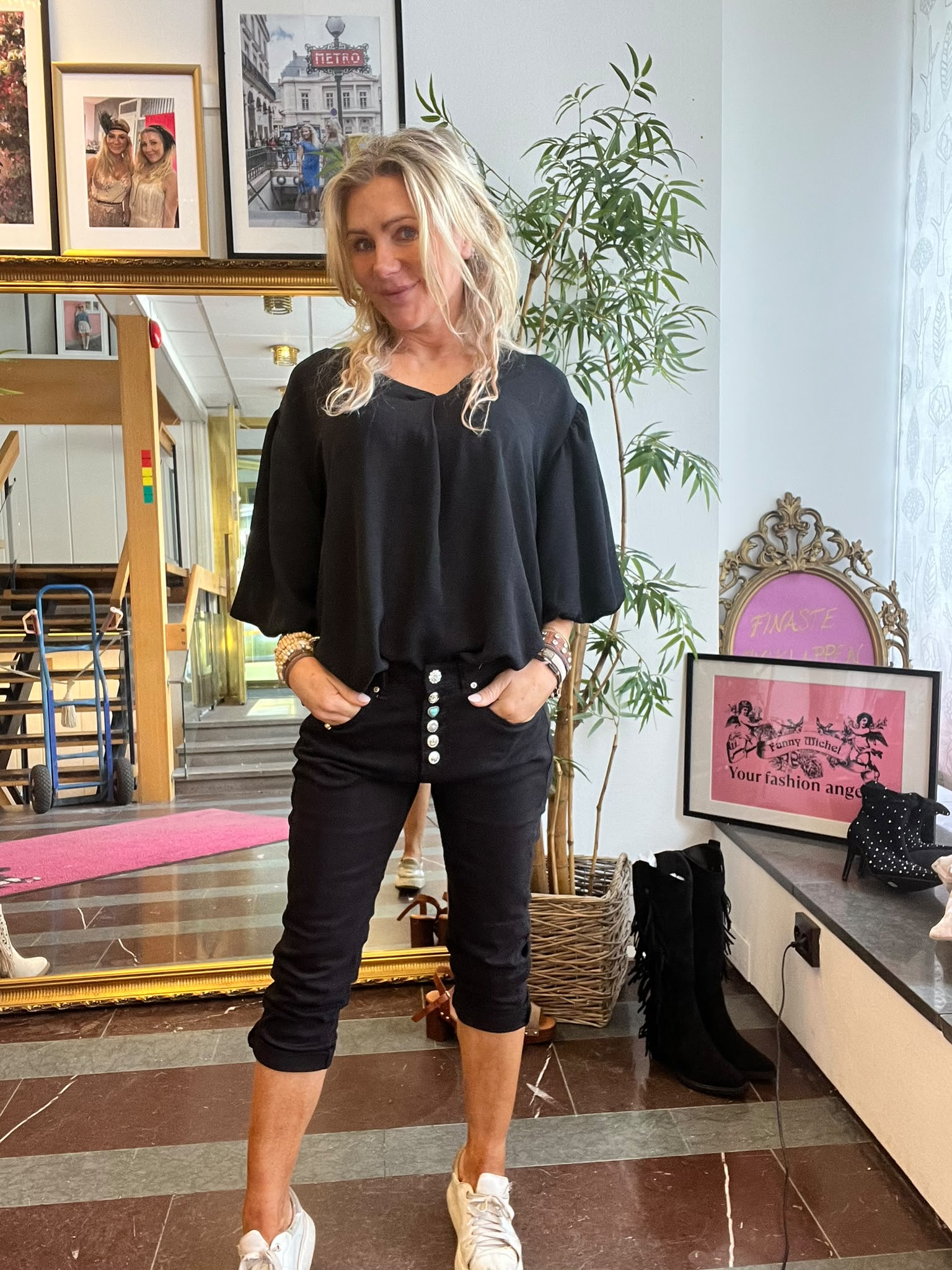 Kvinna i svart blus och svarta byxor står med händerna i fickorna och ler mot kameran i ett rum med stor guldspegel, inramade foton, grön växt och en skylt med texten “Your fashion angel”.
