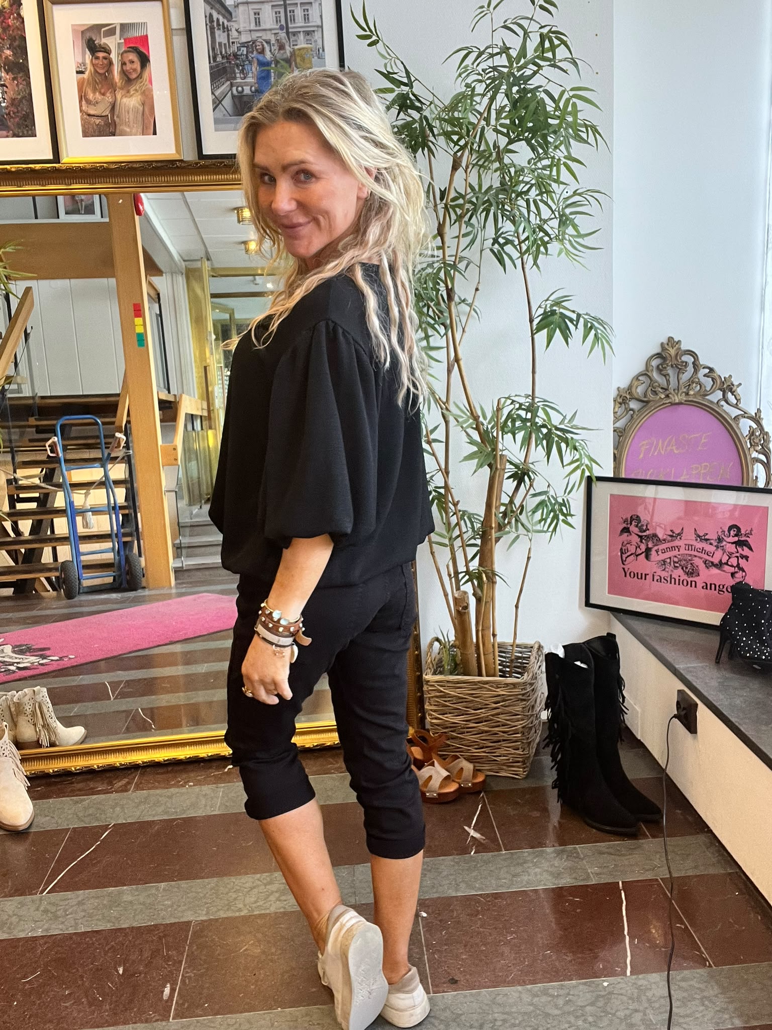 Blond kvinna i svart outfit står och vänder sig mot kameran med ett leende, i en butiksmiljö med spegel, skor och en hög växt. Skylt/affisch med text: “Fanny Michel” och “Your fashion angel”.