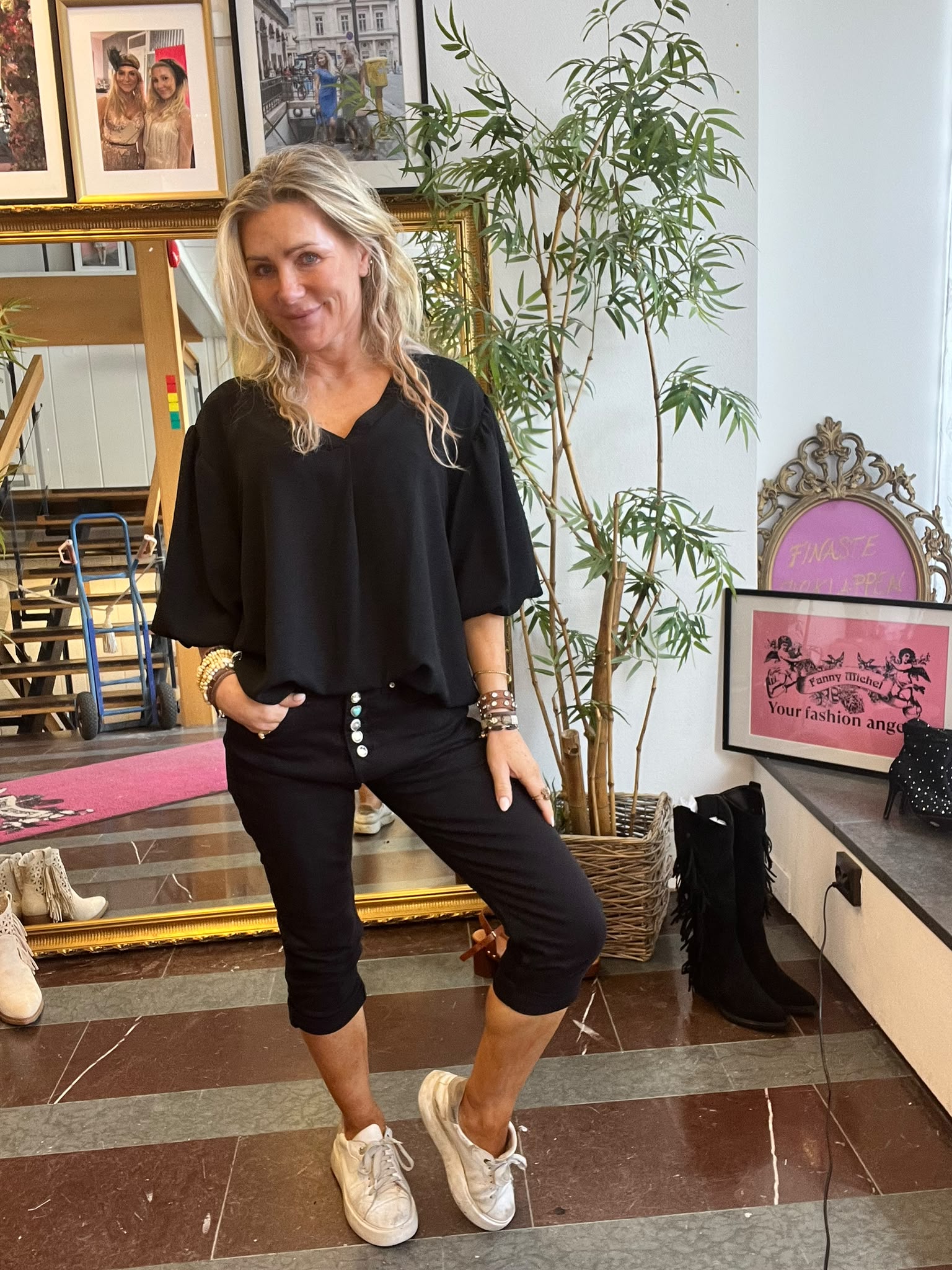 Blond kvinna poserar leende med en hand i fickan, klädd i svart blus och byxor samt vita sneakers. Hon står i en butik/studio med spegel, skoföremål, växt och inramad affisch med texten: “Fanny Michel Your fashion angel”.