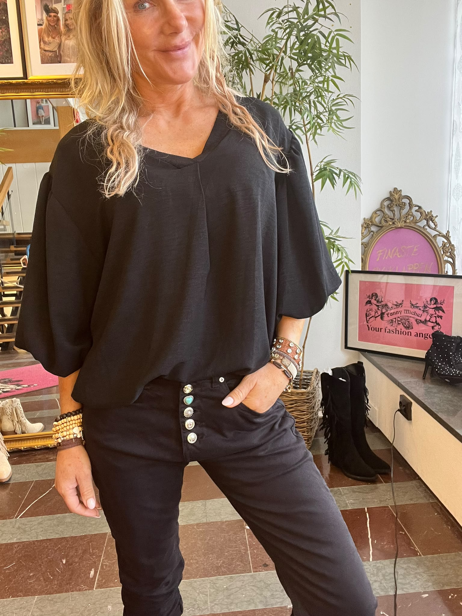 Kvinna med långt blont hår poserar inomhus med handen i fickan, klädd i svart topp och svarta byxor med dekorativa knappar. I bakgrunden syns en växt och en rosa skylt med texten “FANNY MICHEL Your fashion angel”.