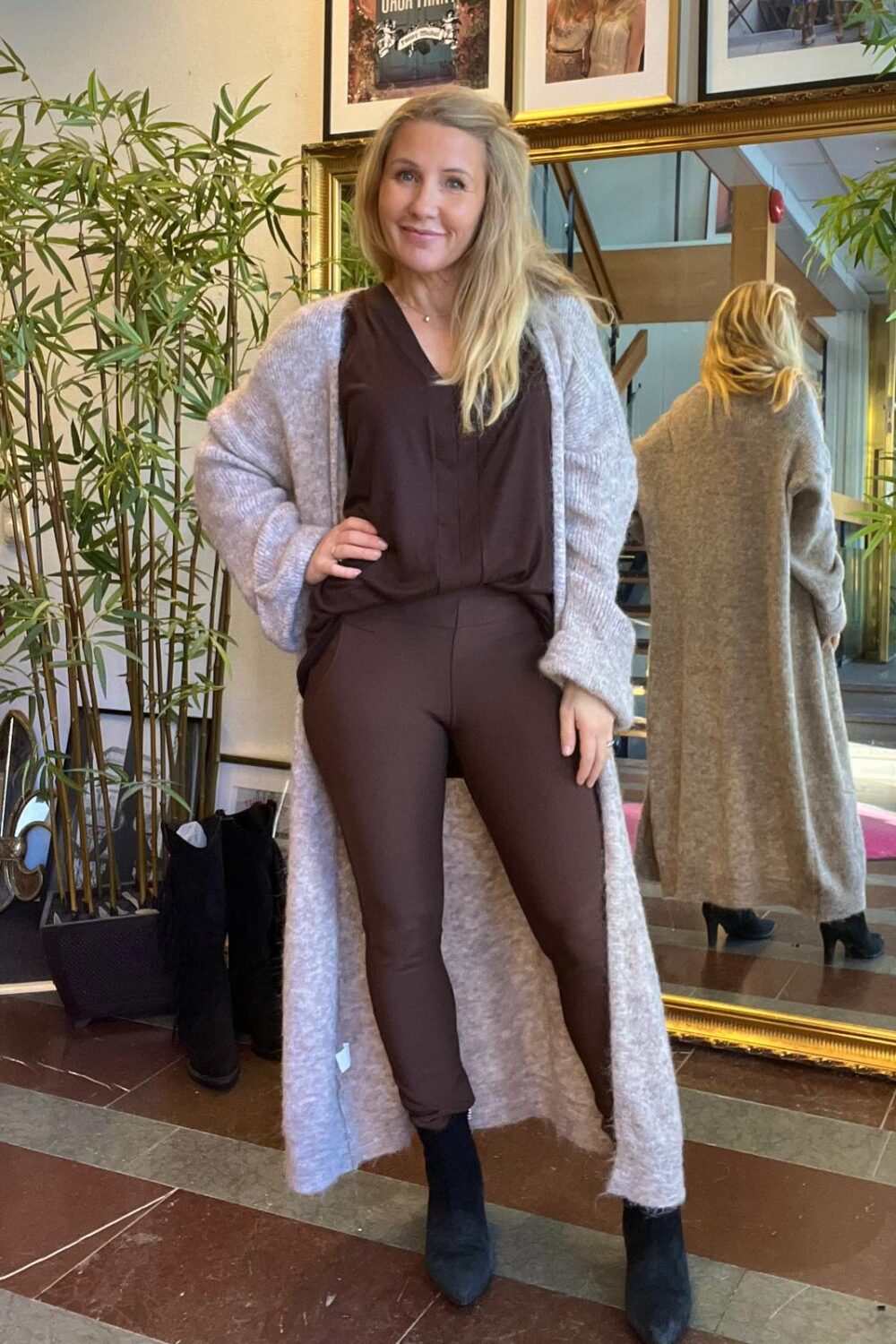 Kvinna med långt blont hår poserar med handen på höften i en hall, klädd i brun outfit och lång grå stickad kofta. Bakom henne speglar en stor guldramsspegel hennes rygg; bambuplantor och tavlor syns.