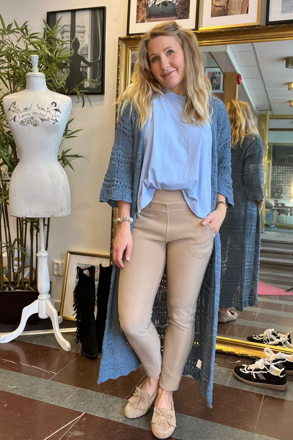 Kvinna poserar inomhus med handen i fickan och visar beige leggings med hög midja, vit topp och lång blå spetskofta. Bakom syns guldramsspegel, klänningsdocka med texten ”Fanny Michel” och växter.