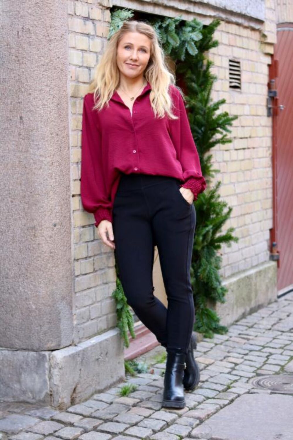 Kvinna i vinröd blus och svarta leggings står lutad mot en tegelvägg med handen i fickan och benen korsade. Hon bär svarta ankelboots och poserar på kullerstensgata vid en grön växt.