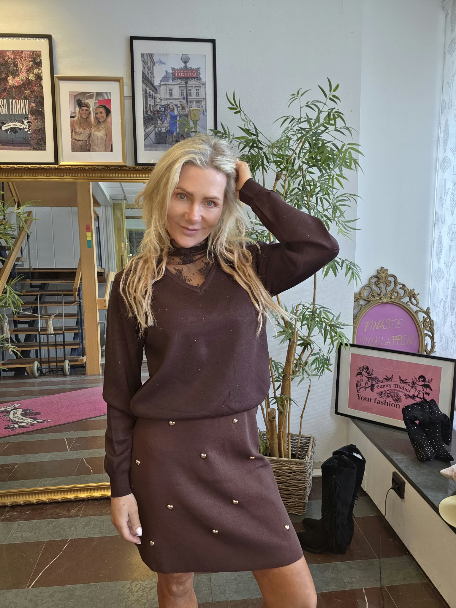 Vaaleahiuksinen nainen poseeraa hymyillen, käsi pään takana, ruskeassa mekossa. Taustalla suuri peili, kehystettyjä kuvia ja tekstejä “METRO” sekä “Fanny Michel Your fashion”, sekä viherkasvi ja koristeellinen tuoli.
