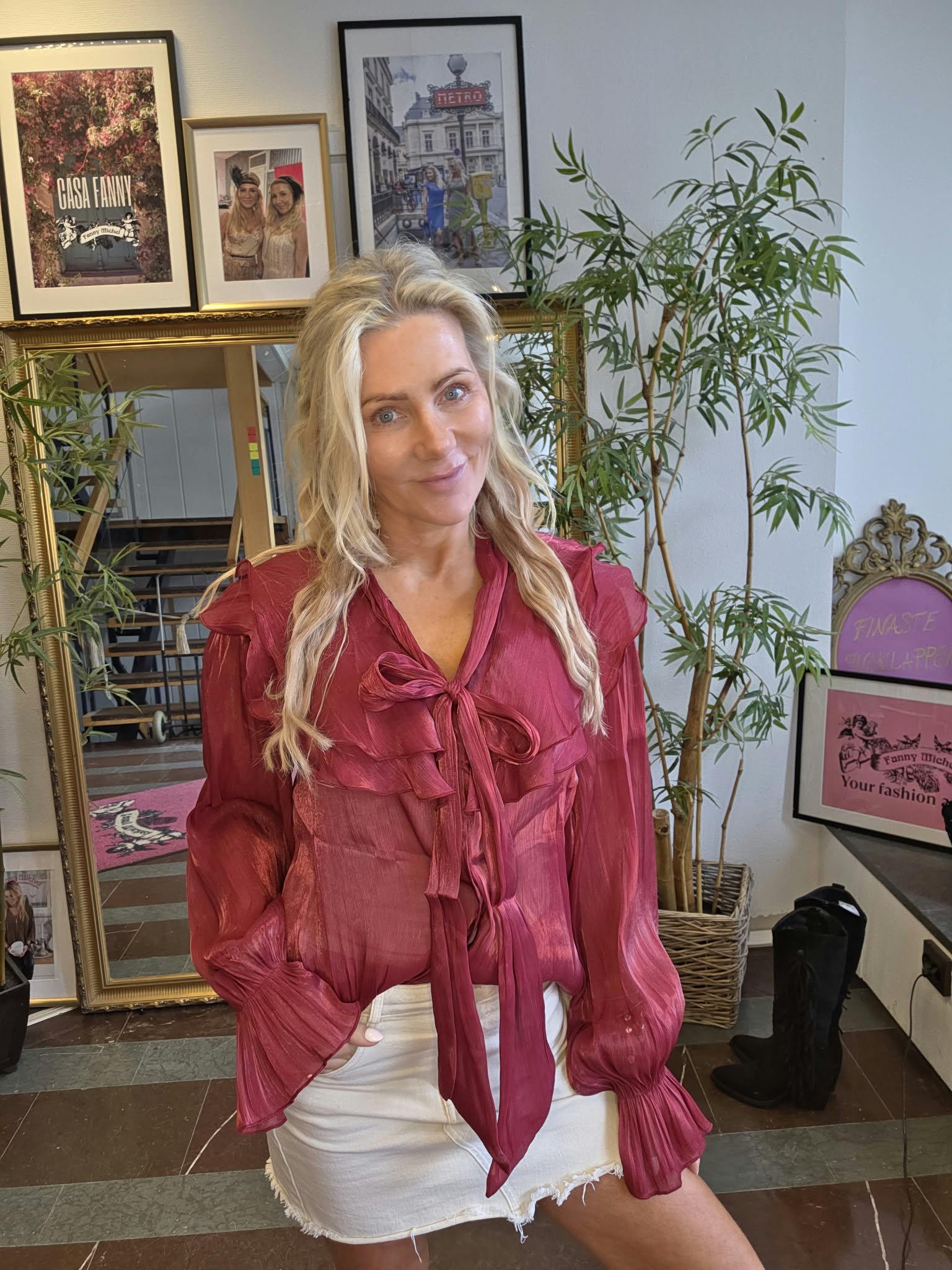 Leende kvinna i röd, rosettknuten blus och vit kjol poserar inomhus framför en spegel och gröna krukväxter. På väggen bakom syns inramade bilder med texten “CASA FANNY” och “METRO”.