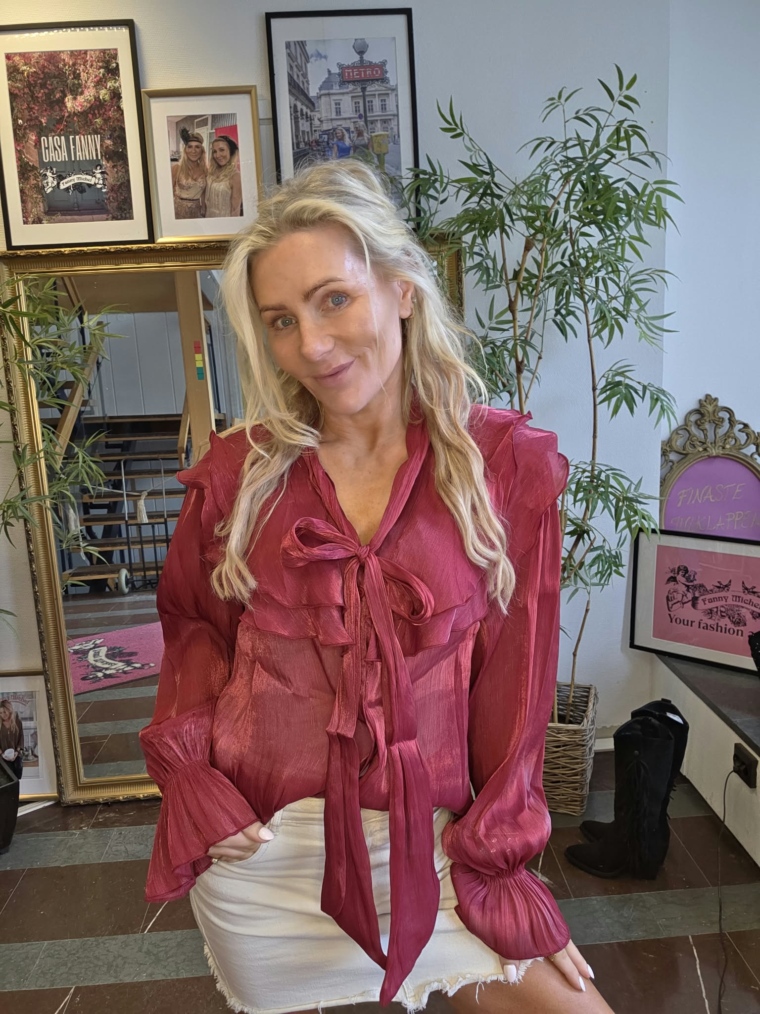 Blond kvinna i vinröd blus med stor rosett lutar sig fram och ler mot kameran i ett rum med spegel, växter och inramade posters. Synlig text: “CASA FANNY”, “METRO”, “FANNY MICHEL”, “Your fashion”.