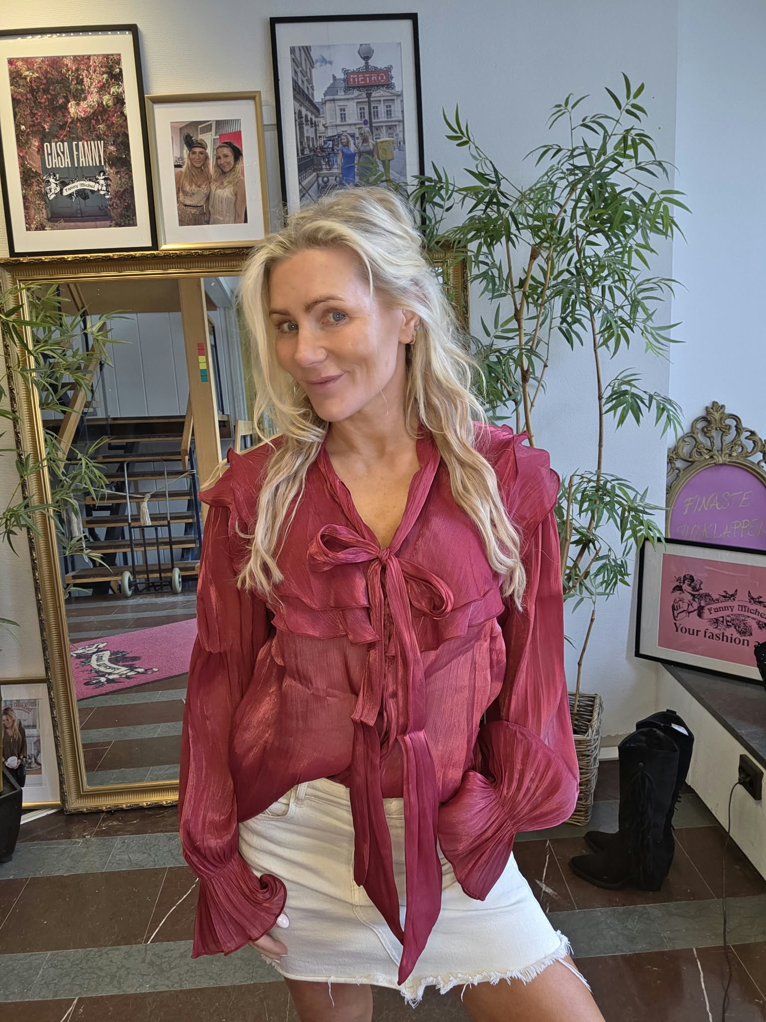 Blond kvinna står och ler mot kameran, klädd i vinröd blus med rosett och vit kjol. Bakom henne syns spegel, växter och inramade bilder med texten “CASA FANNY” och “METRO”.