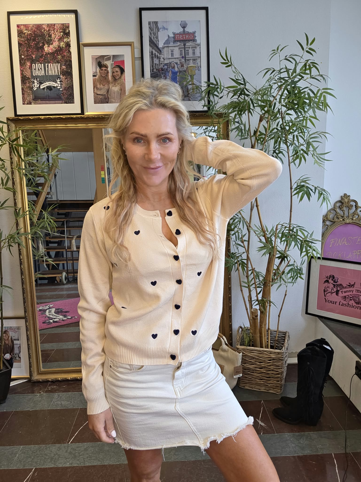 Blond kvinna poserar med ena handen bakom huvudet och ler mot kameran, iklädd beige kofta med mörka hjärtknappar och vit jeanskjol. Hon står i ett rum med spegel, växter och inramade posters, bland annat “CASA FANNY” och “METRO”.
