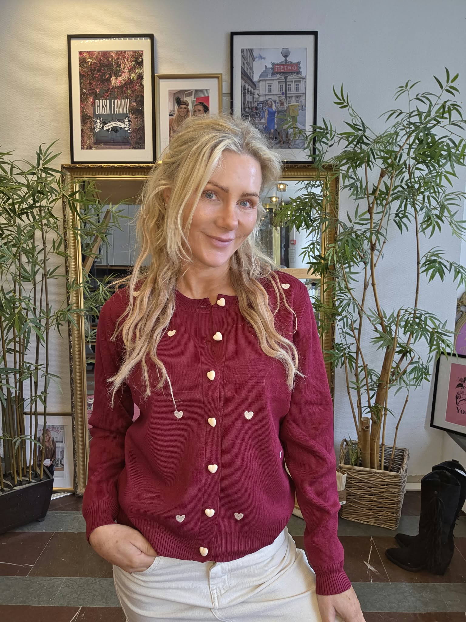 Blond kvinna ler mot kameran och poserar inomhus med handen i fickan. Hon bär vinröd kofta med hjärtknappar och vita byxor, framför en spegel, gröna krukväxter och inramade tavlor med texten ”CASA FANNY” och ”METRO”.
