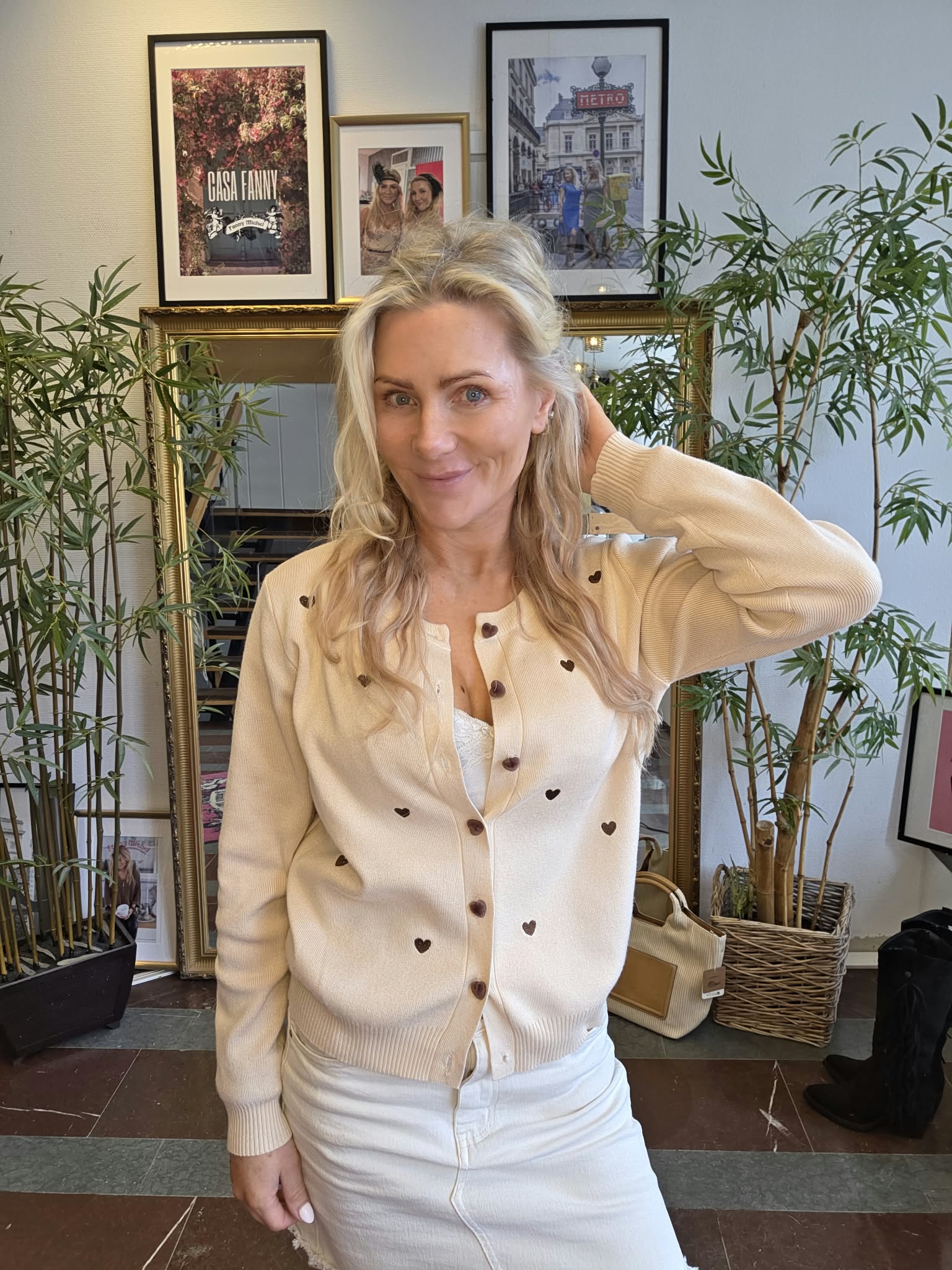 Leende blond kvinna poserar med ena handen bakom huvudet, klädd i beige kofta med små hjärtan och vita byxor. Hon står framför en spegel, omgiven av gröna krukväxter och inramade posters med texten ”CASA FANNY” och ”METRO”.