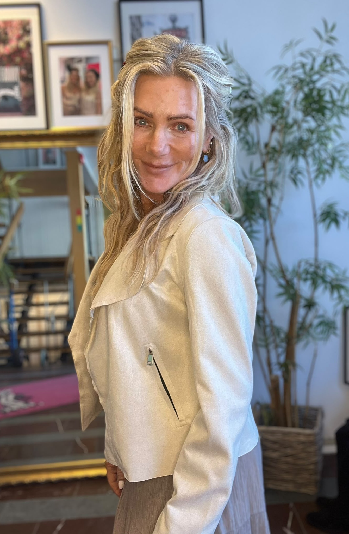Leende blond kvinna står i profil och vänder blicken mot kameran, klädd i ljus kavaj med dragkedjeficka och blå örhängen. I bakgrunden syns inramade foton, en spegel och en grön växt.