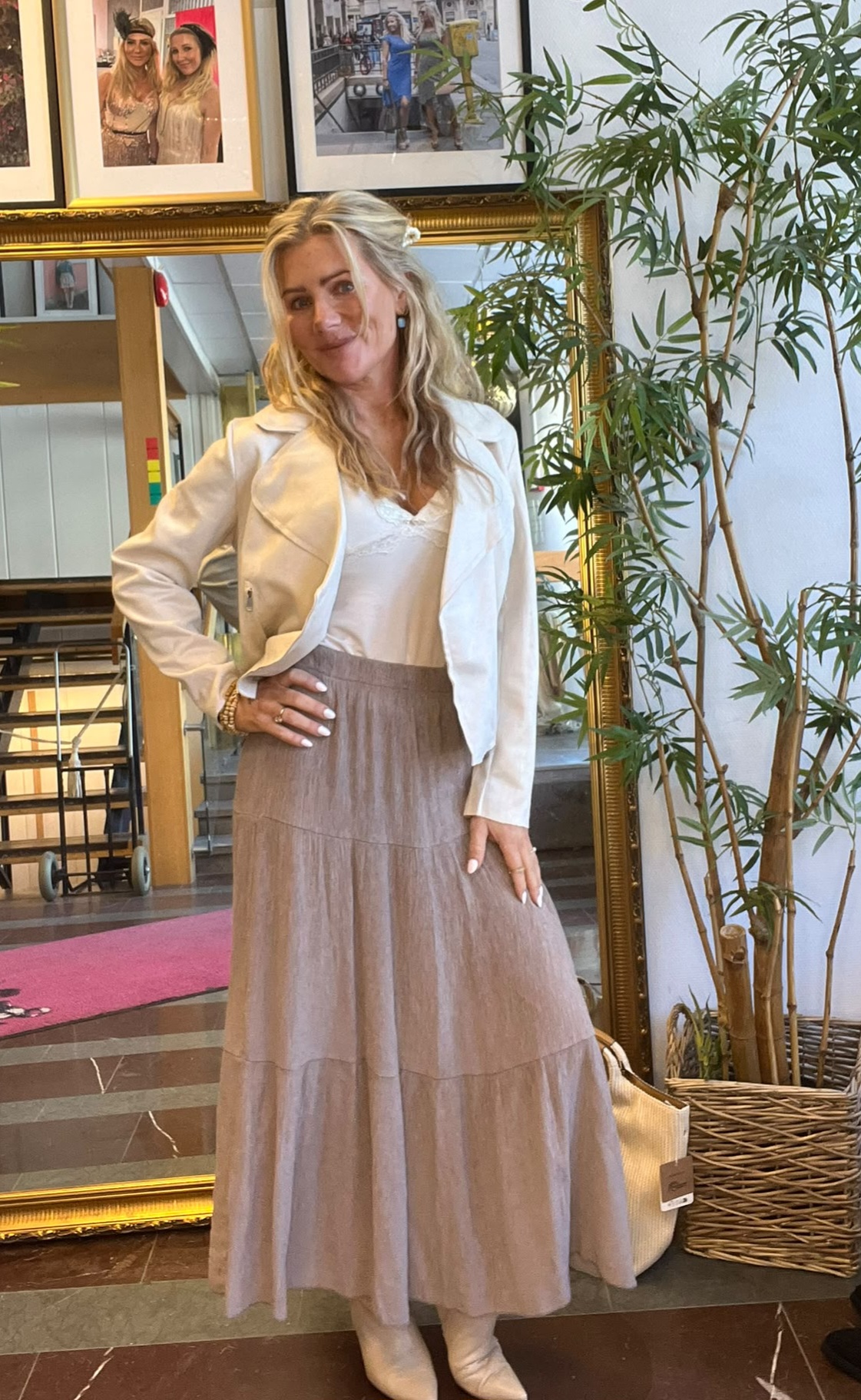Blond kvinna står och poserar med handen på höften framför en stor guldramsspegel, klädd i vit jacka, vit topp och lång beige kjol med ljusa stövlar. I bakgrunden syns inramade foton och en hög krukväxt.