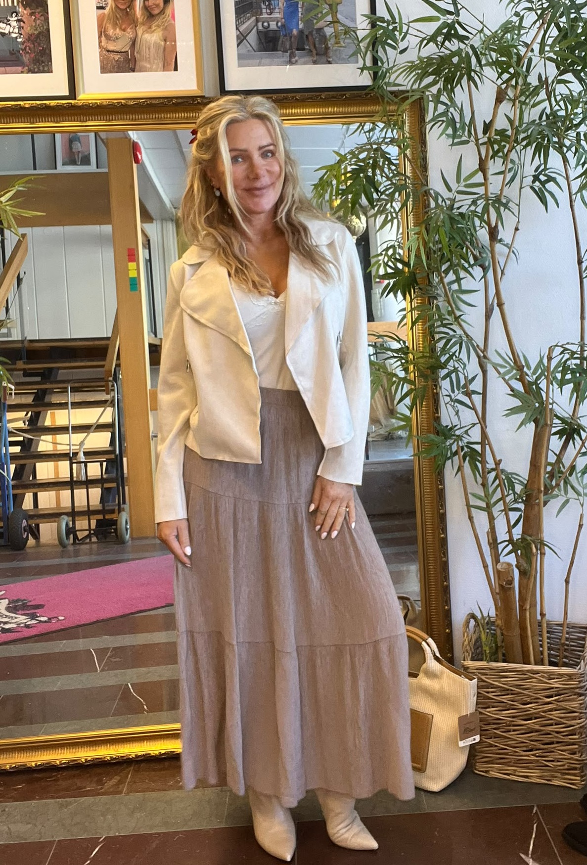 Leende kvinna med långt blont hår står och poserar inomhus framför en stor guldramsspegel, iklädd vit jacka, lång grå kjol och ljusa stövlar; bakom syns inramade foton, en stor krukväxt och en vagn.