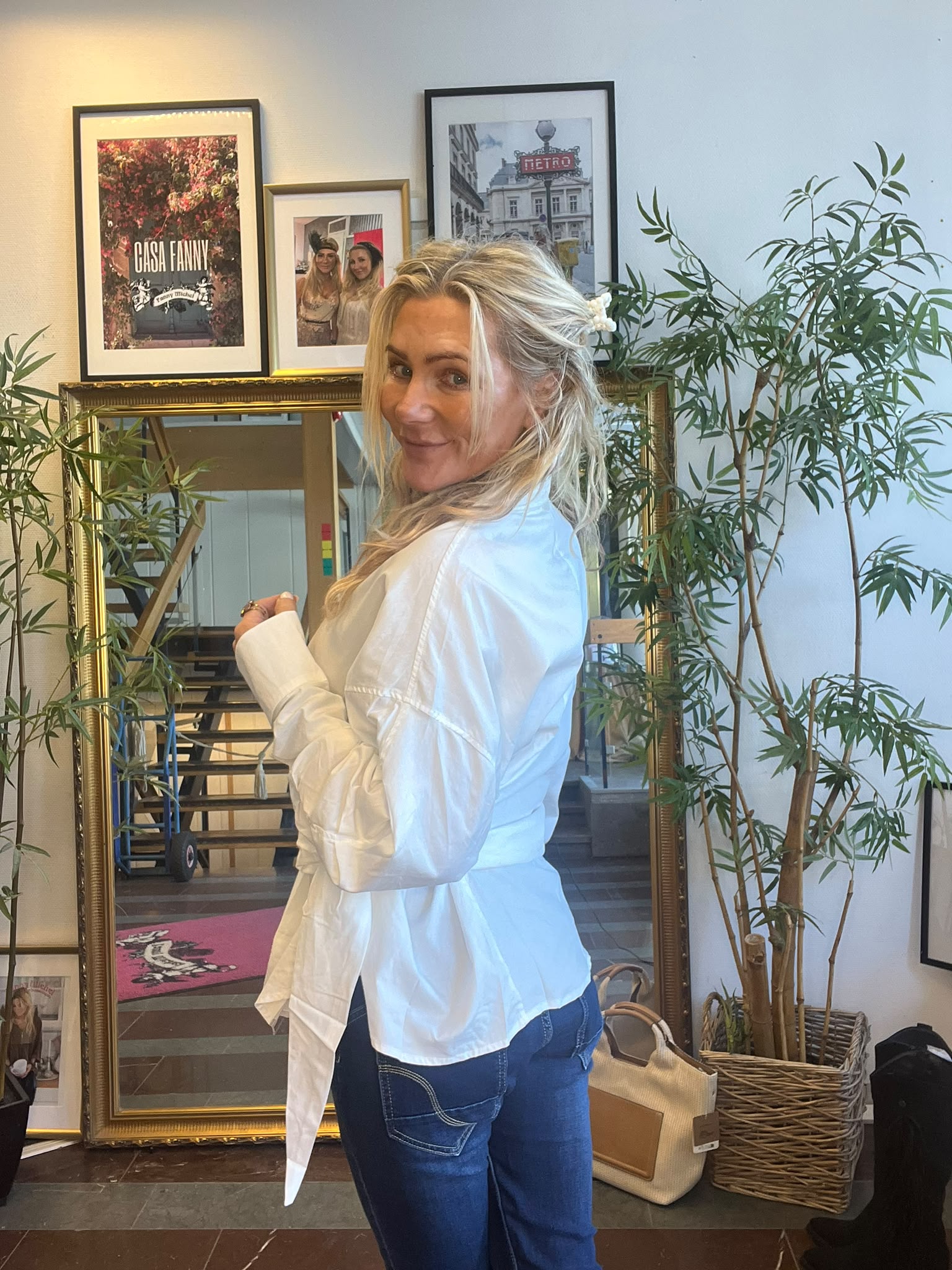 Blond kvinna i vit skjorta och jeans vänder sig om och ler mot kameran i ett rum med stor guldspegel, gröna krukväxter och inramade tavlor på väggen med texten “CASA FANNY” och “METRO”.