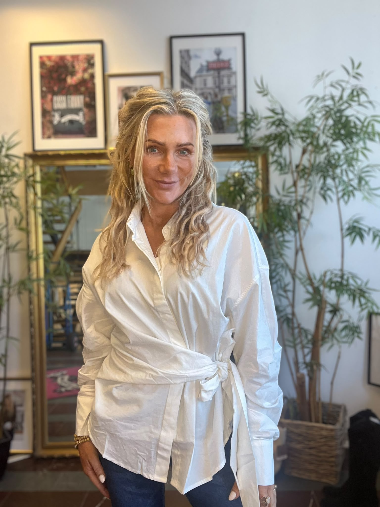 Blond kvinna i vit knuten skjorta och jeans står och ler mot kameran. I bakgrunden syns en bokhylla med inramade konstbilder ovanför samt stora gröna växter i ett ljust rum.