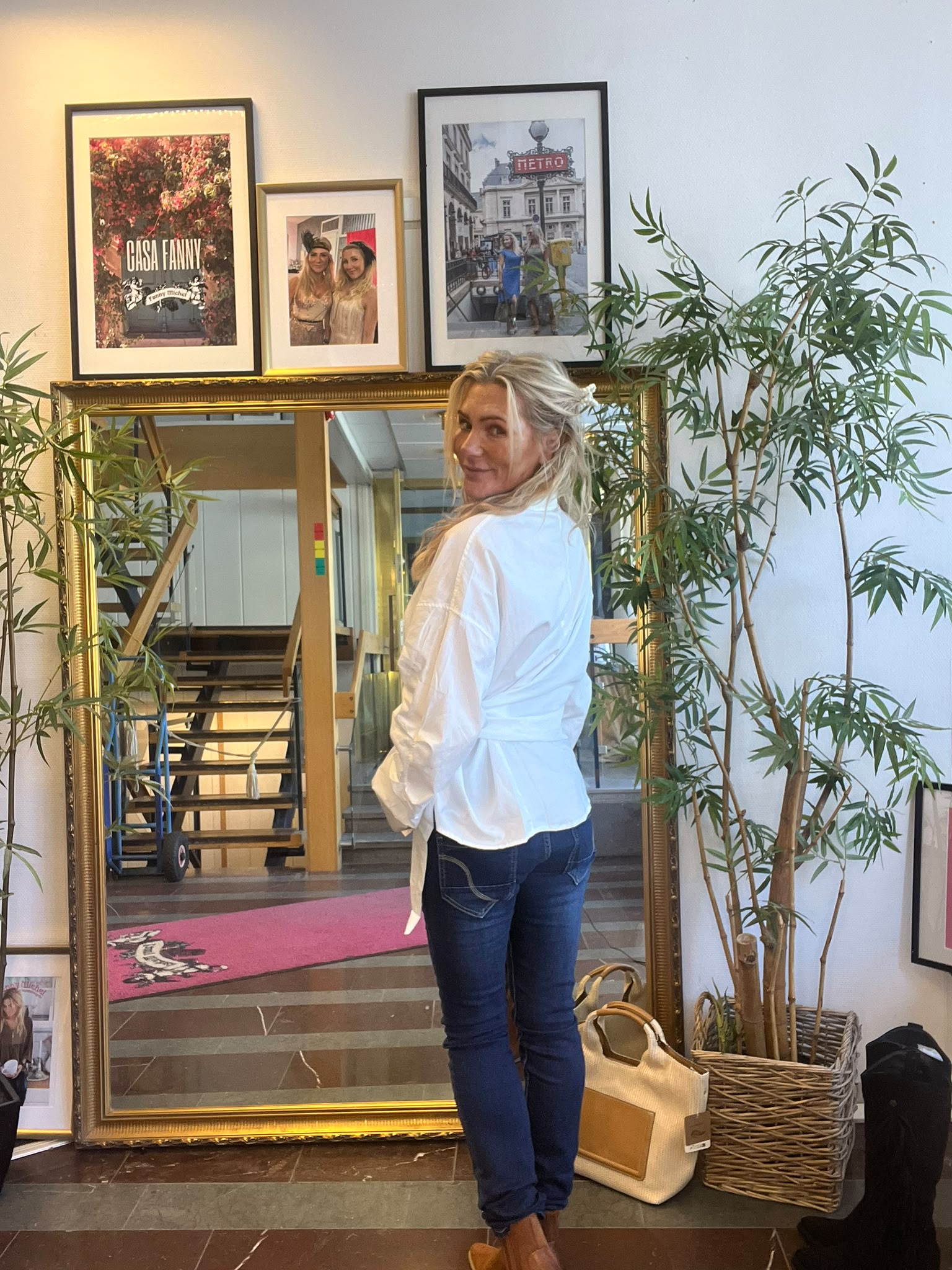 Kvinna i vit blus och jeans står framför en stor guldramsspegel och vänder sig leende mot kameran. Rummet har gröna växter och inramade bilder ovanför, med texten “CASA FANNY” och “METRO”.