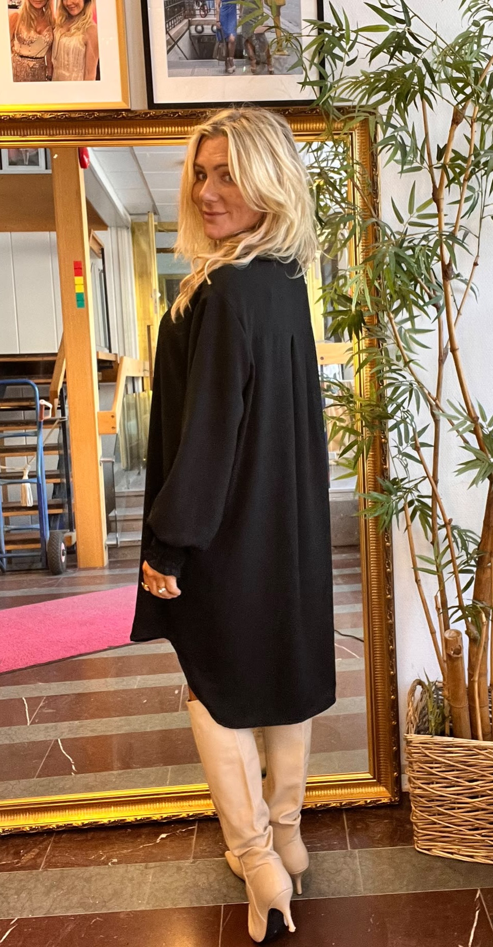 Blond kvinna i svart, löst sittande klänning och ljusa knähöga stövlar står vriden och tittar över axeln mot kameran. Hon poserar framför en stor guldramsspegel bredvid en hög grön växt och inramade foton.