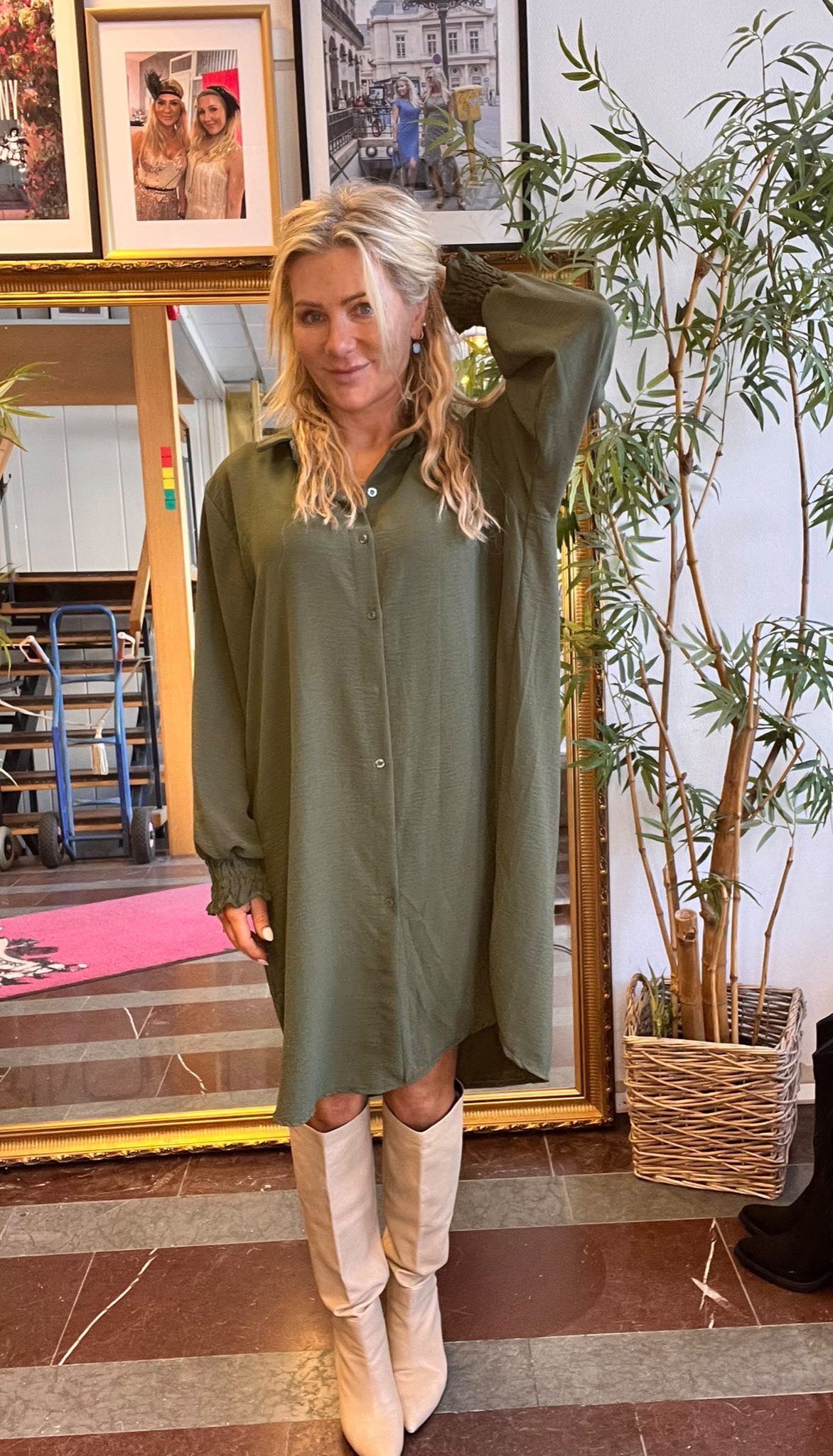 Blond kvinna i grön, lång skjortklänning och vita knähöga stövlar poserar med ena handen bakom huvudet. Hon står inomhus framför en stor guldramsspegel, med inramade foton på väggen och en hög krukväxt bredvid.