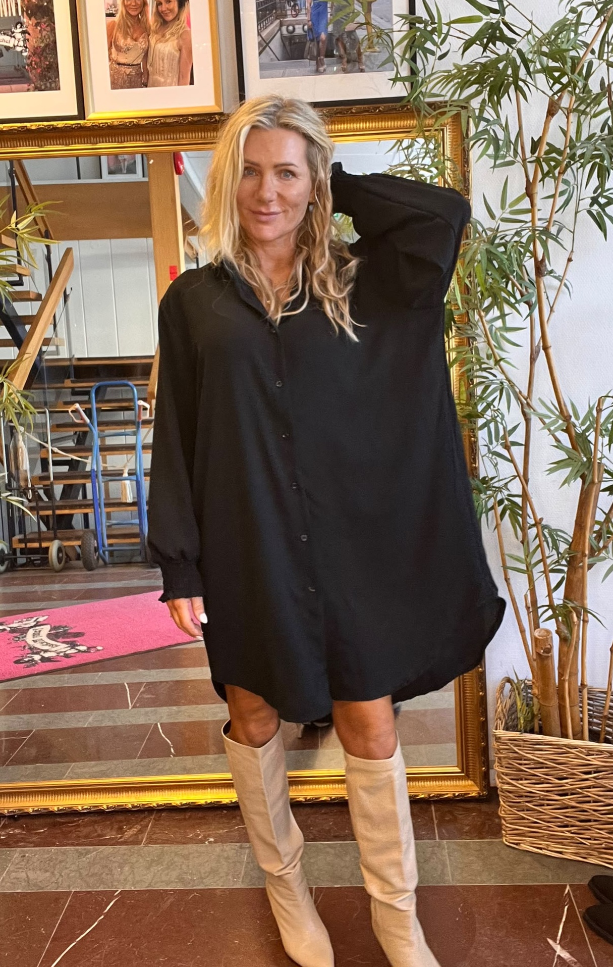 Blond kvinna poserar framför en stor guldramsspegel, med ena handen bakom huvudet. Hon bär en oversized svart skjortklänning och ljusa knähöga stövlar, omgiven av en hög krukväxt och inramade foton på väggen.