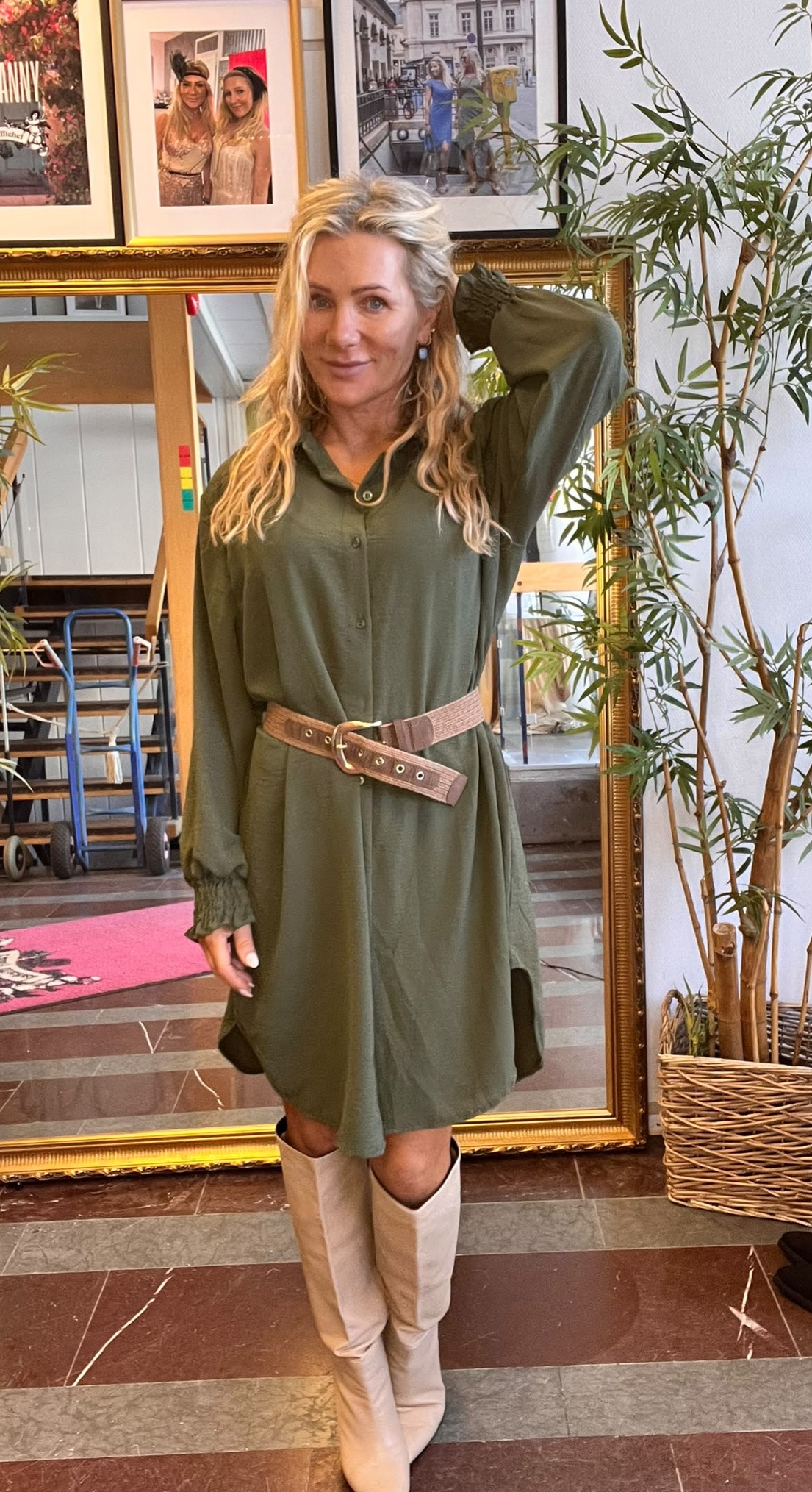Kvinna med långt blont hår poserar med ena handen bakom huvudet, klädd i grön skjortklänning med bredt skärp och ljusa knähöga stövlar. Hon står framför en stor guldramsspegel, med växt och inramade foton på väggen.