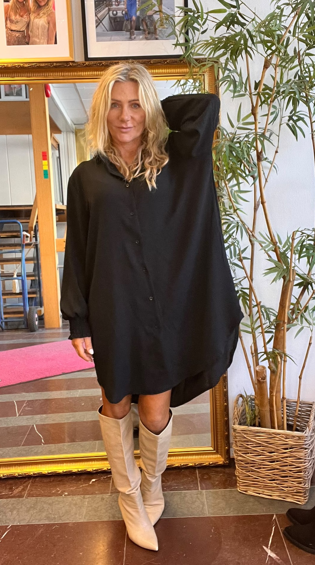 Blond kvinna står och poserar med ena armen bakom huvudet, klädd i en svart, lös skjortklänning och ljusa knähöga stövlar. Hon står framför en stor guldramsspegel bredvid en hög grön växt inomhus.