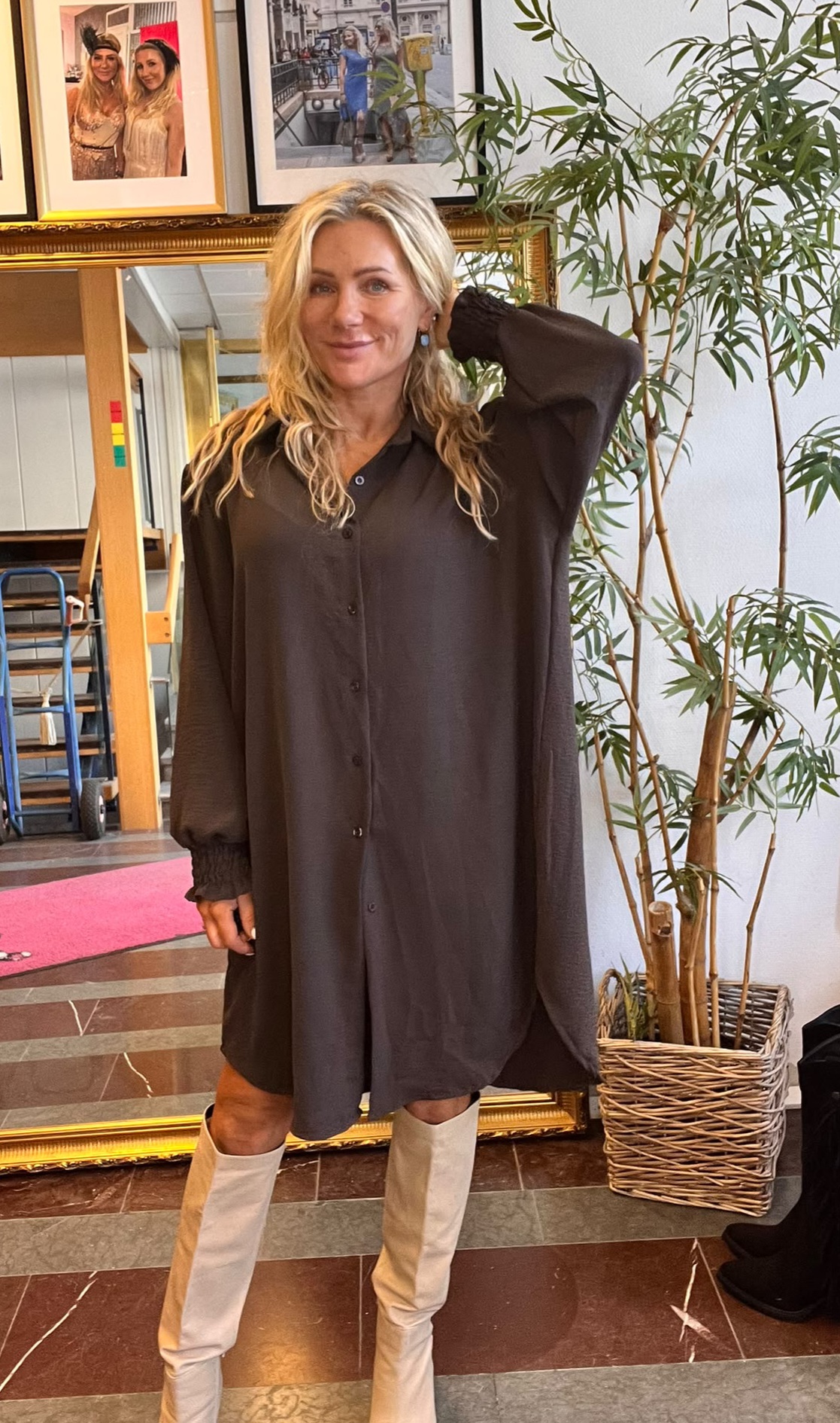 Blond kvinna står och poserar med ena handen bakom huvudet, klädd i en mörk skjortklänning och vita knähöga stövlar. Hon är inomhus bredvid en hög grön växt, med inramade foton på väggen bakom.