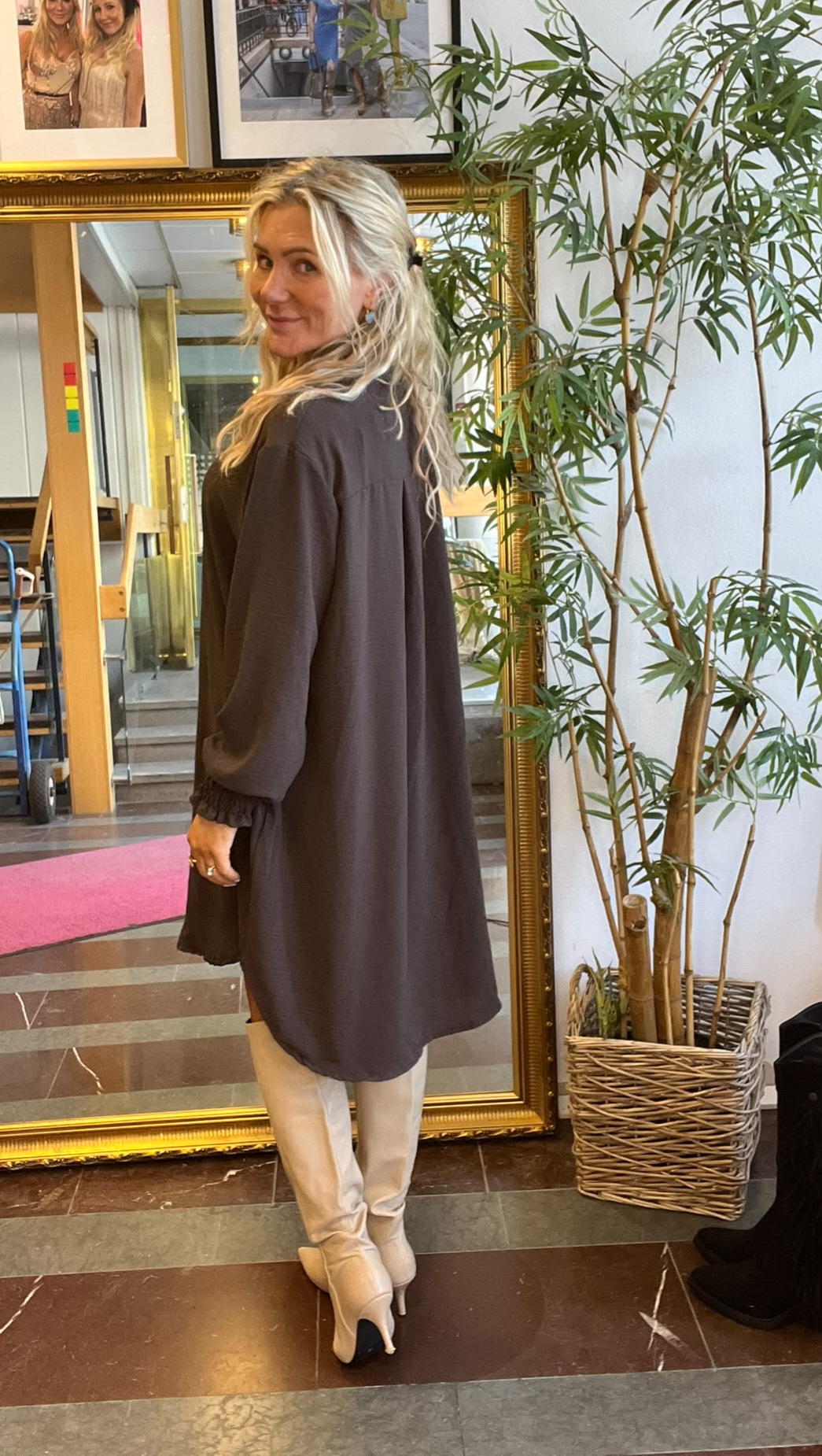 Blond kvinna i mörk, knälång klänning och ljusa stövlar står framför en stor guldramsspegel och vänder sig leende mot kameran. Inomhusmiljö med inramade foton på väggen och en hög grön krukväxt bredvid.