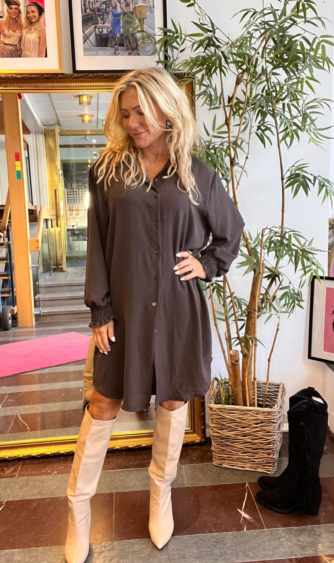 Blond kvinna poserar med handen på höften i ett rum, klädd i en mörk skjortklänning och beige knähöga stövlar. Hon står framför en stor guldramsspegel, med en hög grön krukväxt bredvid.