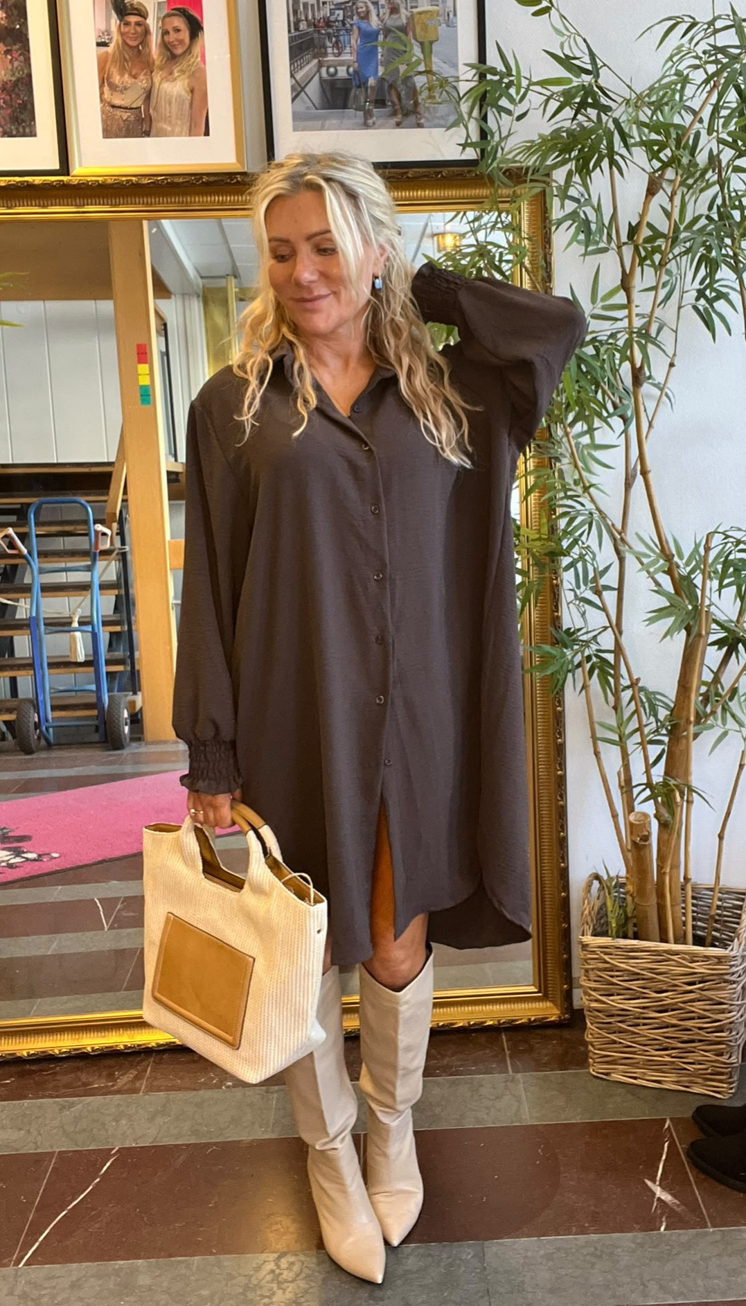 Blond kvinna poserar inomhus med ena handen bakom huvudet och en ljus handväska i den andra. Hon bär mörk skjortklänning och vita knähöga stövlar, framför en guldramsspegel, växt och inramade foton på väggen.