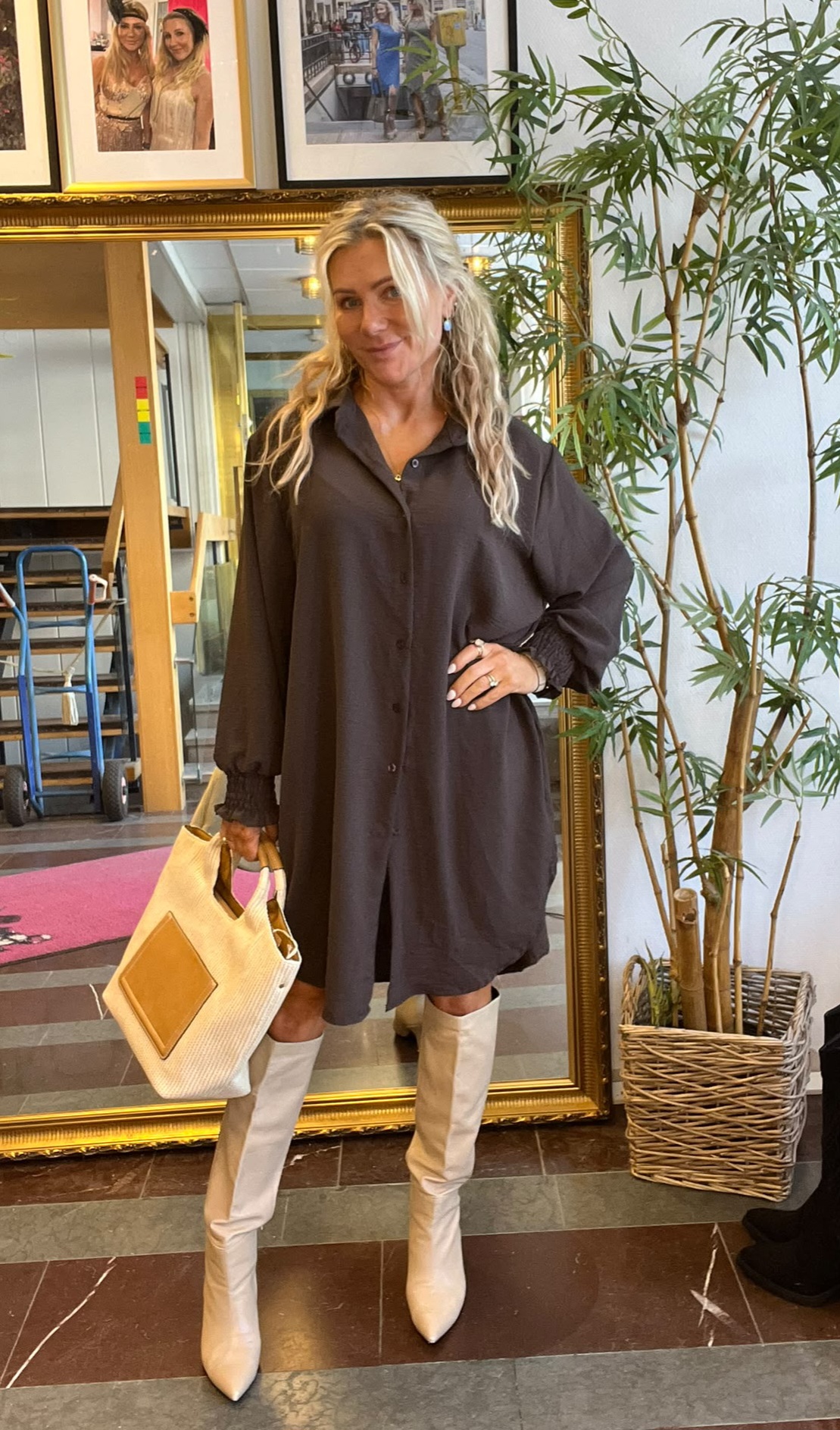 Kvinna med långt blont hår poserar med handen på höften och håller en beige väska. Hon bär mörk skjortklänning och vita knähöga stövlar, framför en stor guldramsspegel med inramade foton och en hög krukväxt bredvid.