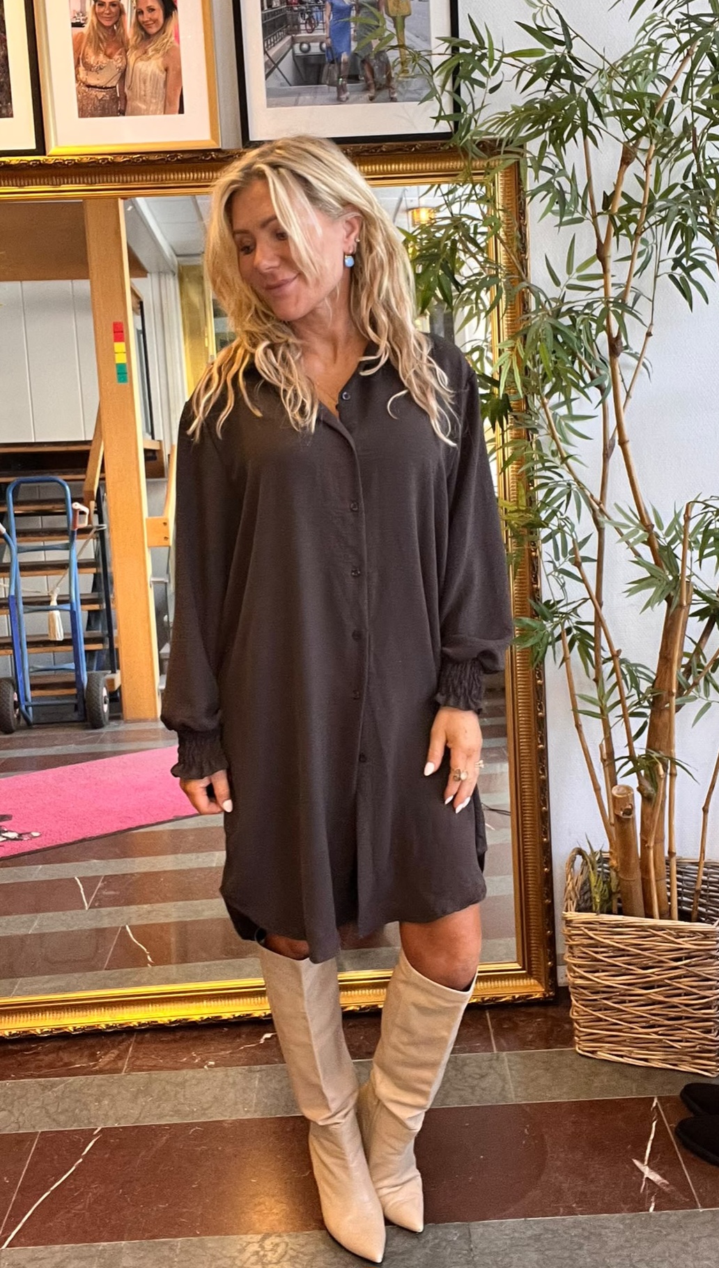 Blond kvinna står och poserar med blicken nedåt, iklädd svart knäppt skjortklänning och beige knähöga stövlar. Inomhus framför stor guldramsspegel, med bambuliknande krukväxt och inramade foton på väggen.