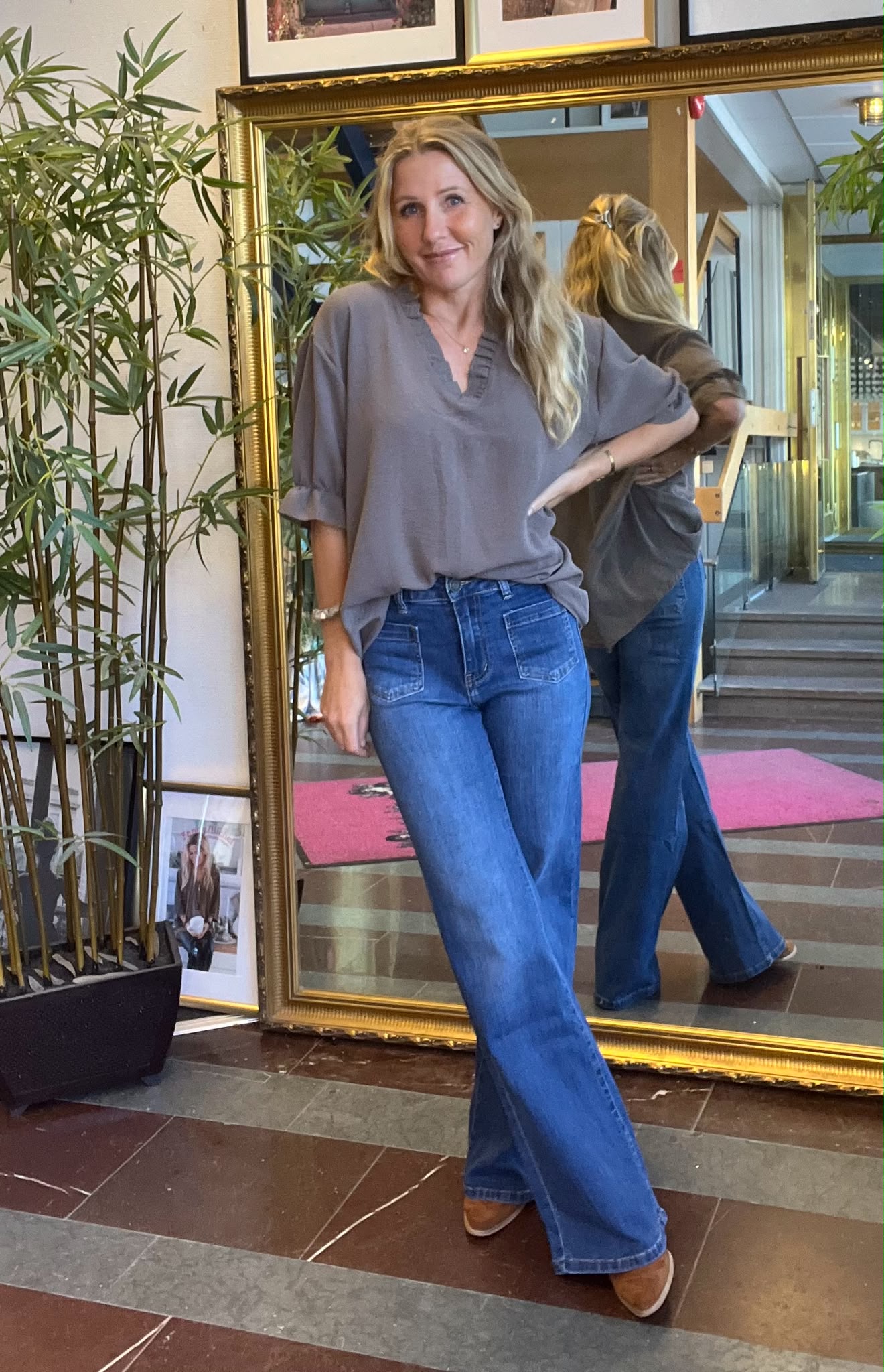 Kvinna med långt blont hår poserar med handen i fickan framför en stor guldramsspegel, iklädd grå blus, blå utsvängda jeans och bruna skor. Spegelbilden syns bakom, med bambuplanta och trappa i bakgrunden.