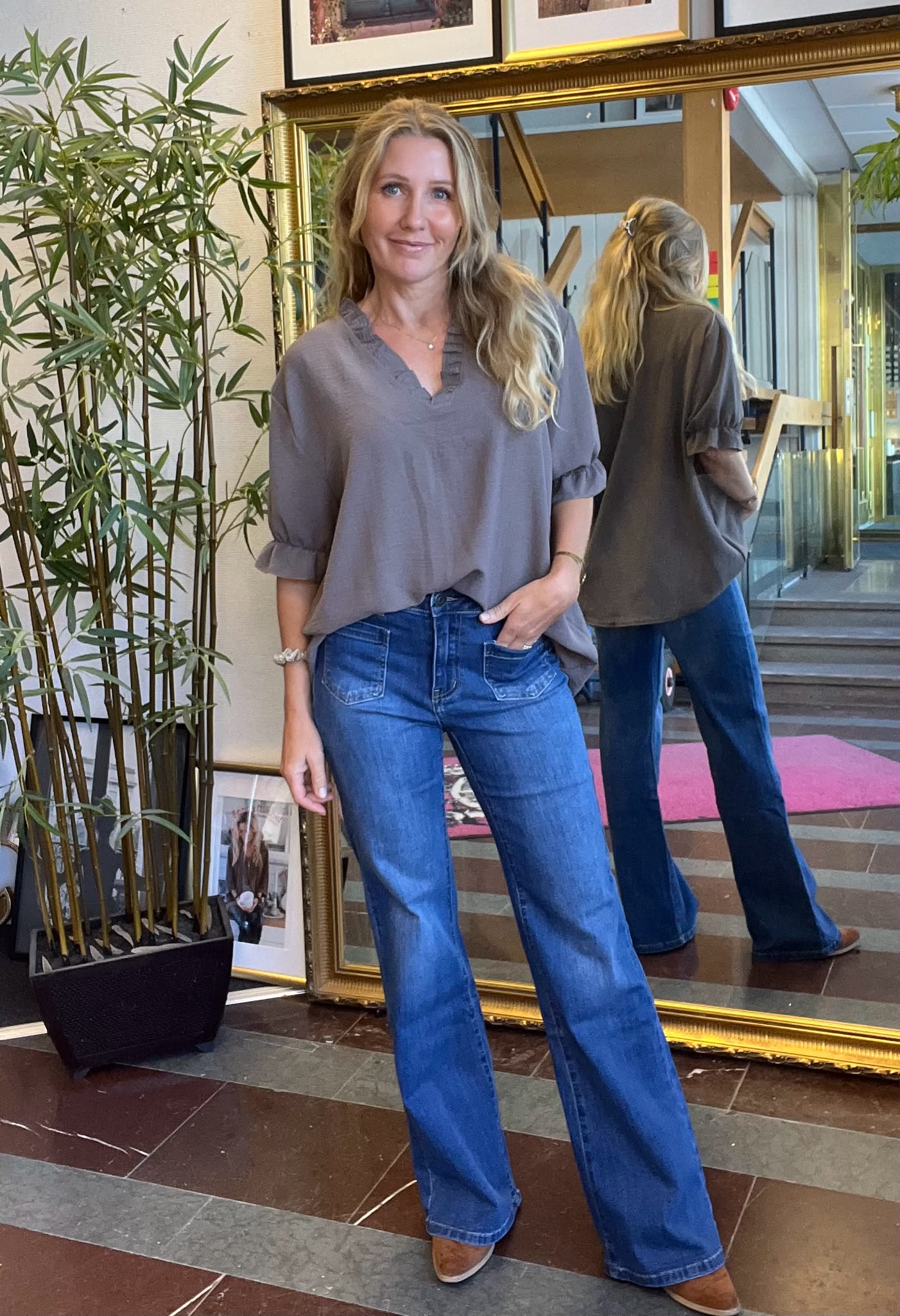Kvinna står och poserar med en hand i jeansfickan och ler mot kameran. Hon bär grå blus, blå bootcut-jeans och bruna skor. Bakom syns hennes spegelbild i en stor guldram, samt en hög grön växt.