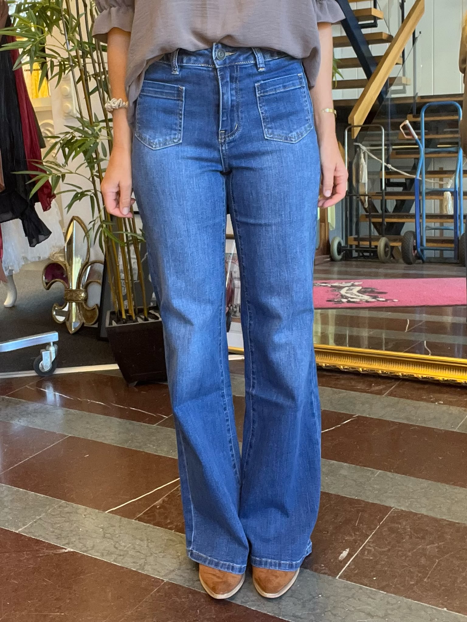 Person står inomhus och visar blå bootcut-jeans med hög midja och stora framfickor, tillsammans med en grå topp och bruna skor. I bakgrunden syns en trappa, klädställning och en stor växt.