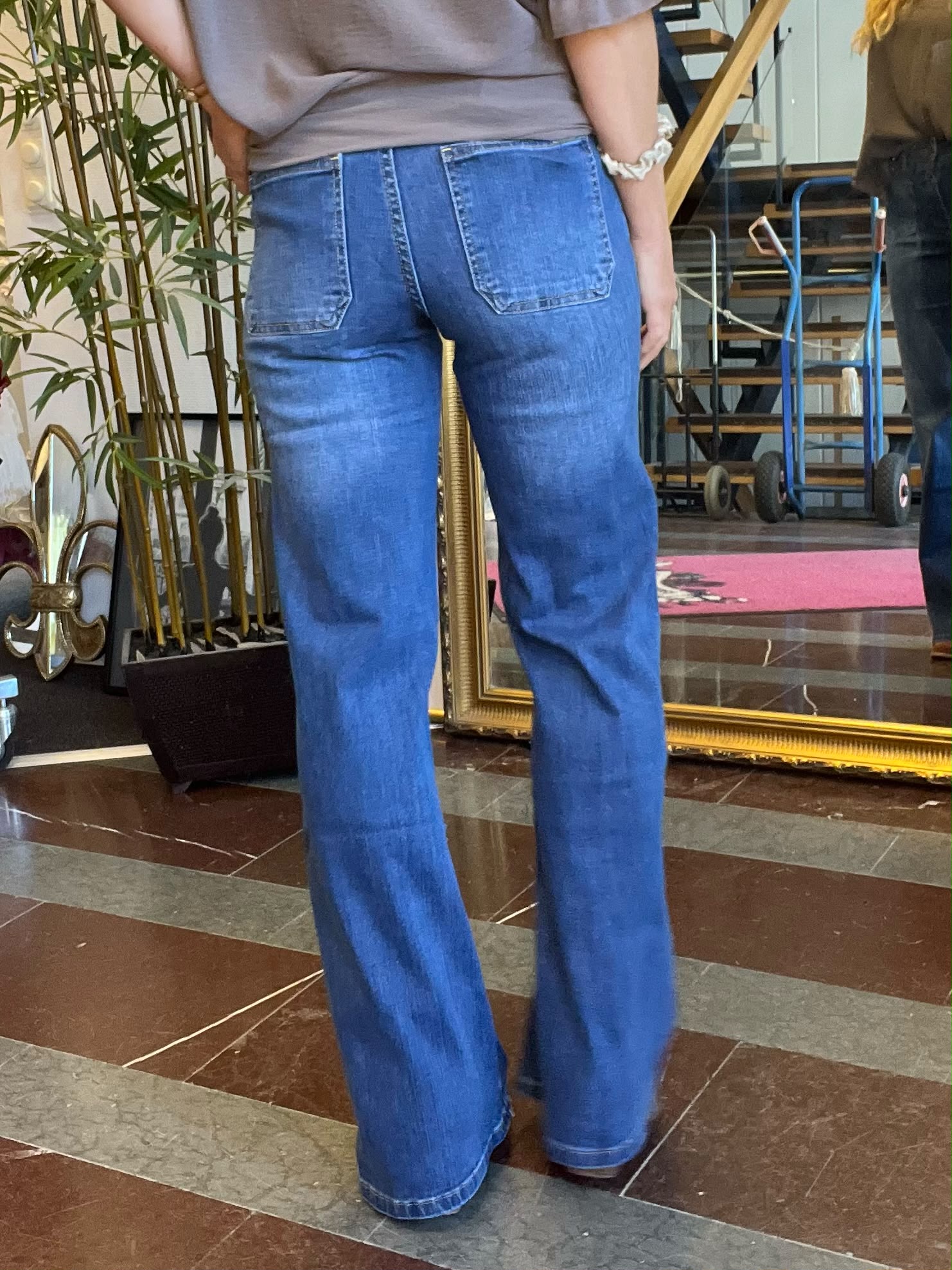 Person i blå bootcut-jeans står med ryggen mot kameran och ena handen i bakfickan. Miljön är en inomhuslobby med stor guldramsspegel, krukväxt och marmorliknande golv; en säckkärra och trappa syns i bakgrunden.